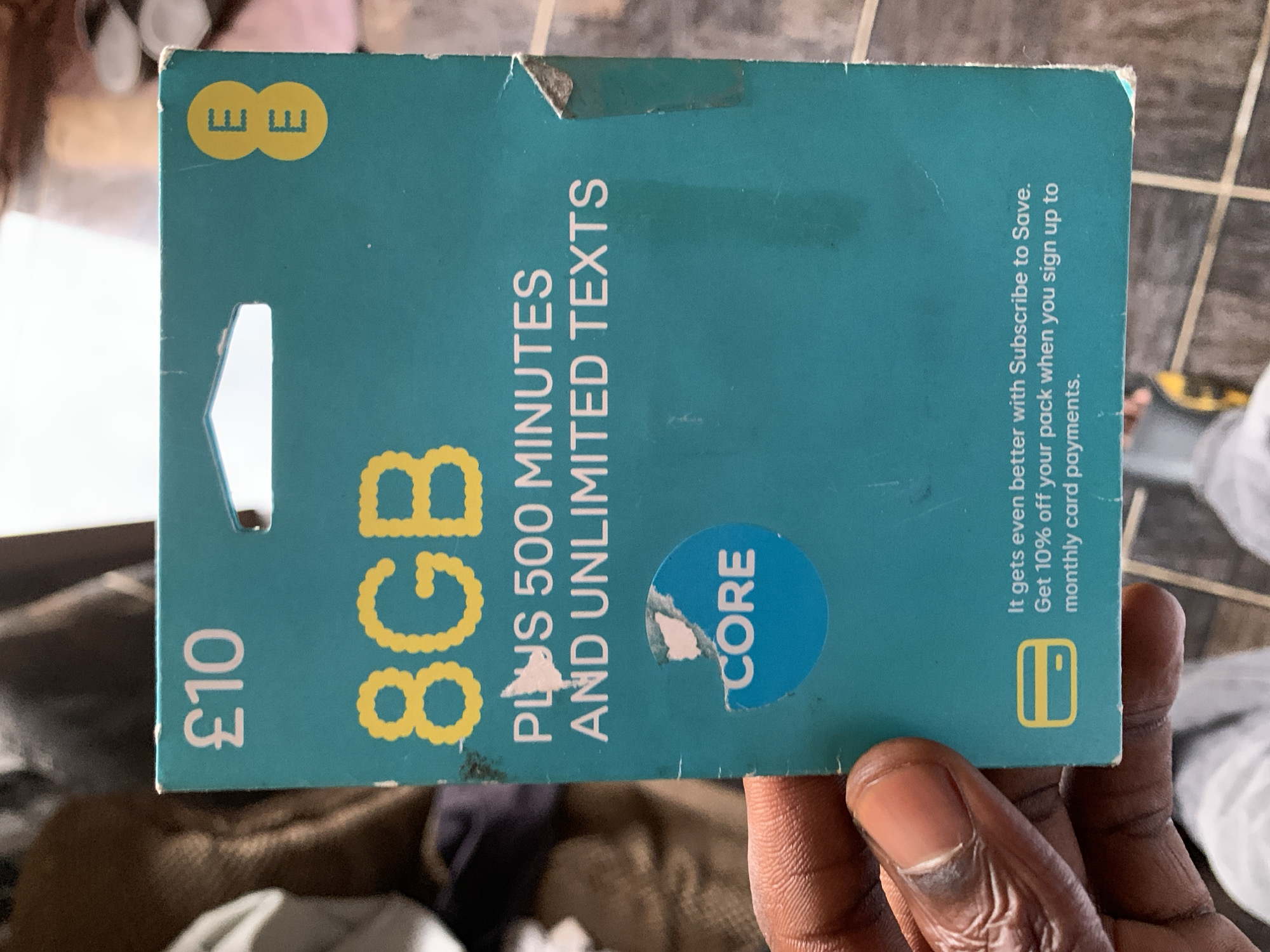 The image shows a close-up of a teal-colored SIM card packaging with text and logos. In the top left corner, "£10" is printed in white. Below this, "8GB" is written in large yellow bubble letters. Further down, "PLUS 500 MINUTES AND UNLIMITED TEXTS" is printed in white text. A circular blue logo with white text reading "CORE" is visible on the left side, partially obscured by a torn white sticker. In the top right corner, two yellow circles with the letter "E" inside each are present, representing the "EE" logo. Near the bottom right, a small yellow icon of a credit card is displayed next to text that reads "It gets even better with Subscribe to Save. Get 10% off your pack when you sign up to monthly card payments." The packaging appears to be held by a person whose hand and fingers are visible on the left side of the image. The background is blurred and appears to be an indoor setting with a tiled floor and some fabric elements. The overall condition of the packaging shows signs of wear and tear, including rips and scuffs. The image is taken from a slightly downward angle.