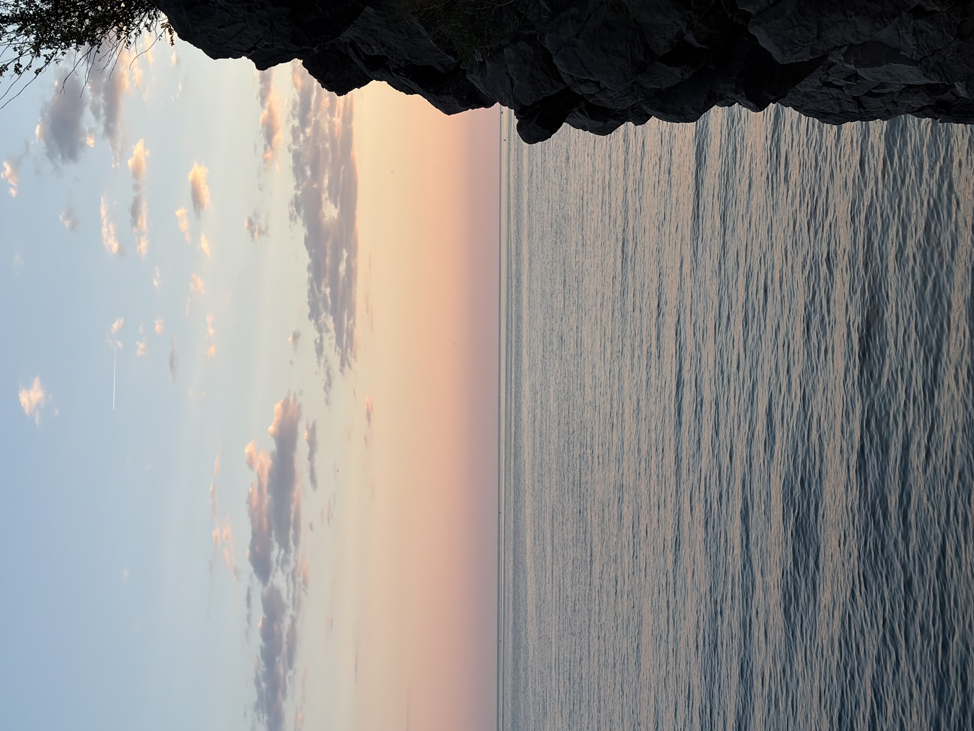 A serene landscape featuring a body of water, possibly the Adriatic Sea, fills the right half of the image, its surface textured with subtle ripples. The left half showcases a sky transitioning from a pale blue to a soft pink-orange hue, dotted with scattered clouds. A rocky overhang, possibly a cliff face, dominates the top of the image. This overhang includes vegetation. The straight line between the water and sky, the color of the sunset, and the cliffs suggest a location along the coast of Budva, Montenegro. No people are visible. A contrail from an aircraft is barely visible in the sky on the left.