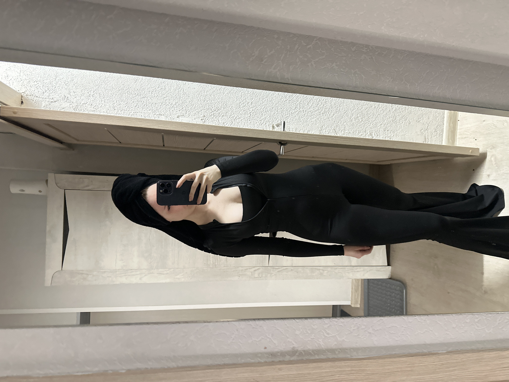 A medium-shot mirror selfie captures a woman in a black, long-sleeved jumpsuit. She holds a smartphone with a black case in her right hand, partially obscuring her face. Her hair is covered by a black towel. The background includes a light wood shelf with a white wall above and below. The location is unknown.