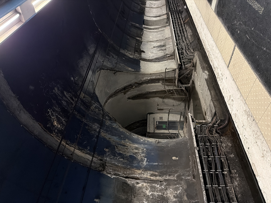This image depicts a section of the abandoned Porte des Lilas - Cinéma station in the Paris Métro system, located in the 19th arrondissement of Paris, France. The scene is an underground subway platform and track area, characterized by severe concrete decay and water damage.
The vaulted ceiling is painted a faded, peeling dark blue, revealing exposed concrete and extensive efflorescence, particularly along the arches and where electrical conduits are mounted. Fluorescent light fixtures illuminate the scene from the upper right. The tunnel wall exhibits prominent arched recesses. In the leftmost arch, a modern, gray metal door with a small window and a green sign is recessed. A rudimentary metal handrail extends from the wall near this door. To the right of this, the concrete wall features vertical ribbing and substantial staining, with another, more open arched structure further along the tunnel.
Adjacent to the platform edge, electrical cables are systematically bundled and routed along the lower tunnel wall within a concrete conduit system. Below this, a dark, water-filled trough runs alongside the track bed. The platform itself is constructed of concrete, with a tactile paving strip of yellow squares visible along its edge in the foreground, adjacent to a dark gray track area. No individuals, actions, or interactions are visible. The environment conveys a sense of disused functionality and significant structural age.