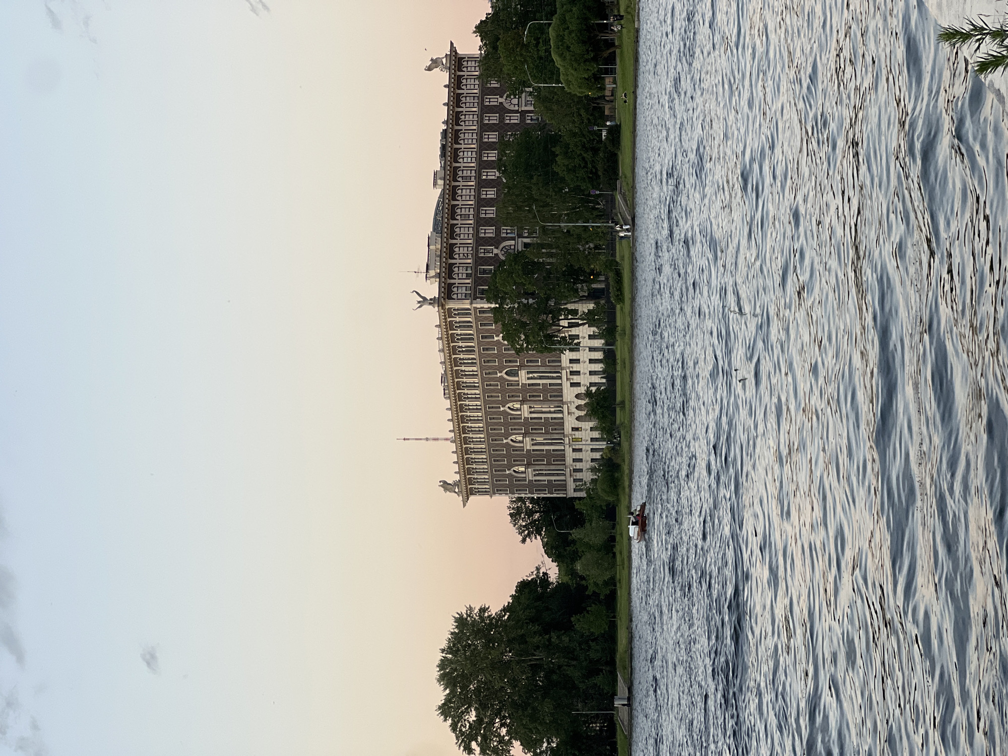 The image shows a wide body of water in the foreground, with ripples reflecting the soft pastel colors of the twilight sky. In the middle ground, a small red boat with two people aboard is navigating the water. Behind the boat, a large, ornate building with a dark facade and numerous windows stands prominently. This building appears to be a historic structure, possibly an apartment building or a government institution, with architectural details such as decorative elements on the roofline. To the left of the building, a large, mature tree with lush green foliage contributes to the serene landscape. The background consists of the sky, transitioning from a pale blue at the top to a warm, peachy hue near the horizon, with scattered clouds. The overall scene suggests a peaceful evening by the water in Saint Petersburg, Russia, likely overlooking the Neva River or one of its tributaries.
