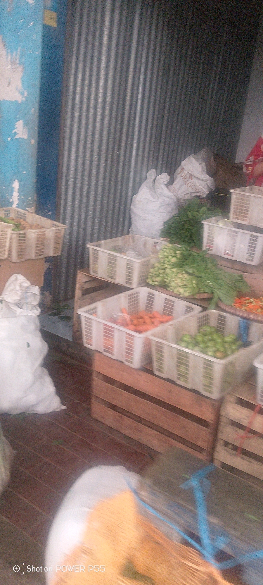 In an outdoor market stall, produce is displayed in white plastic baskets on wooden crates. The baskets contain carrots, leafy green vegetables, and what appear to be small green tomatoes. In the background, a corrugated metal wall creates a textured backdrop, and two large white sacks are visible. To the left, a blue painted wall with peeling paint is present. The ground is paved with reddish-brown bricks. A person's legs and feet, wearing red and white clothing, are visible on the right side of the image. A blurry object, possibly a woven basket with wooden skewers, is in the foreground. The scene is dimly lit.