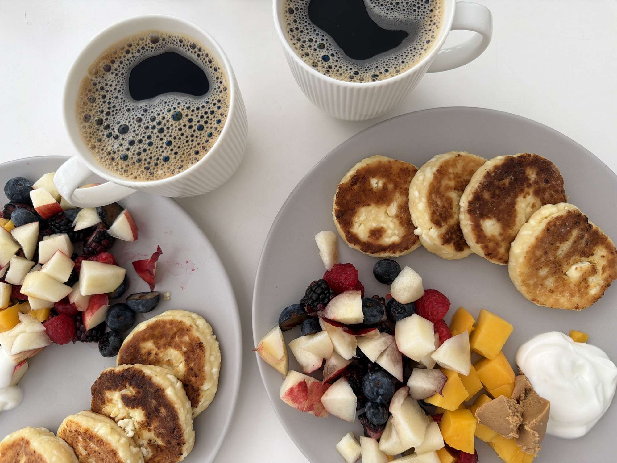 Two plates of syrniki (cheese pancakes) are arranged on a white surface, one above the other. The top plate contains three syrniki and a medley of chopped fruits, including peaches, raspberries, blackberries, and blueberries, along with a dollop of yogurt. The bottom plate holds four syrniki, a pile of diced mango, a dollop of peanut butter, and a swirl of yogurt. Two white mugs of black coffee, each with a foamy surface, are positioned to the right of the plates. The overall scene depicts a breakfast spread. There is no indication of the city of Bat Yam, Israel, in the image or its context.