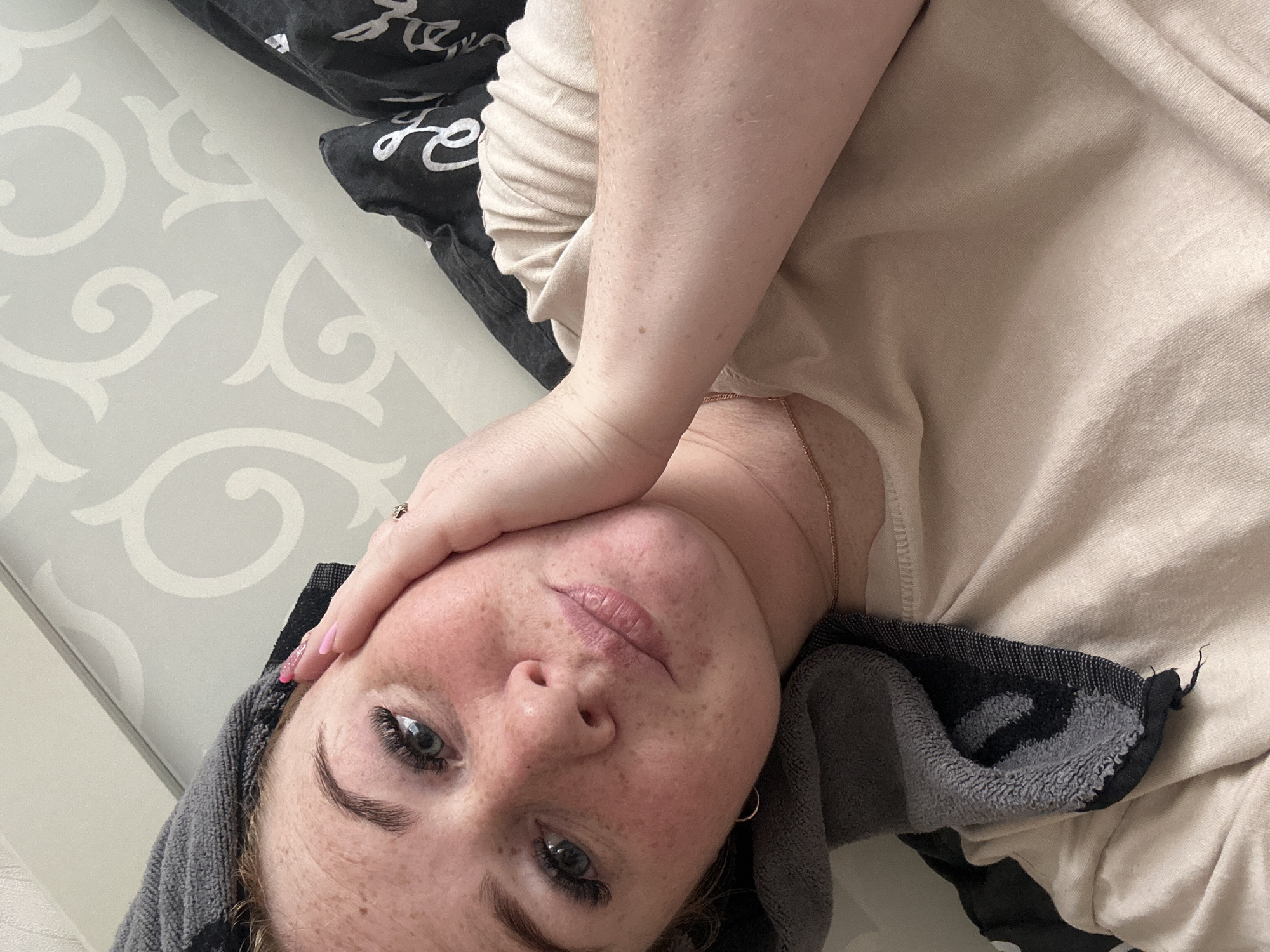A young woman with freckles and blue eyes, wearing a beige t-shirt and a grey towel wrapped around her head, rests her head on her hand. Her nails are painted pink. She is lying in bed, with a patterned wallpaper behind her and a dark pillow visible. The image is taken from a high angle, focusing on her face and upper body. There is no indication of the location being Chaikovsky, Russia.