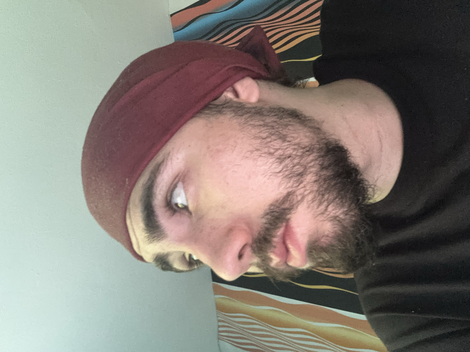 A man is pictured from the shoulders up, looking to his left. He has a beard and is wearing a dark red bandana on his head and a black t-shirt. Behind him is a wall with a striped pattern.