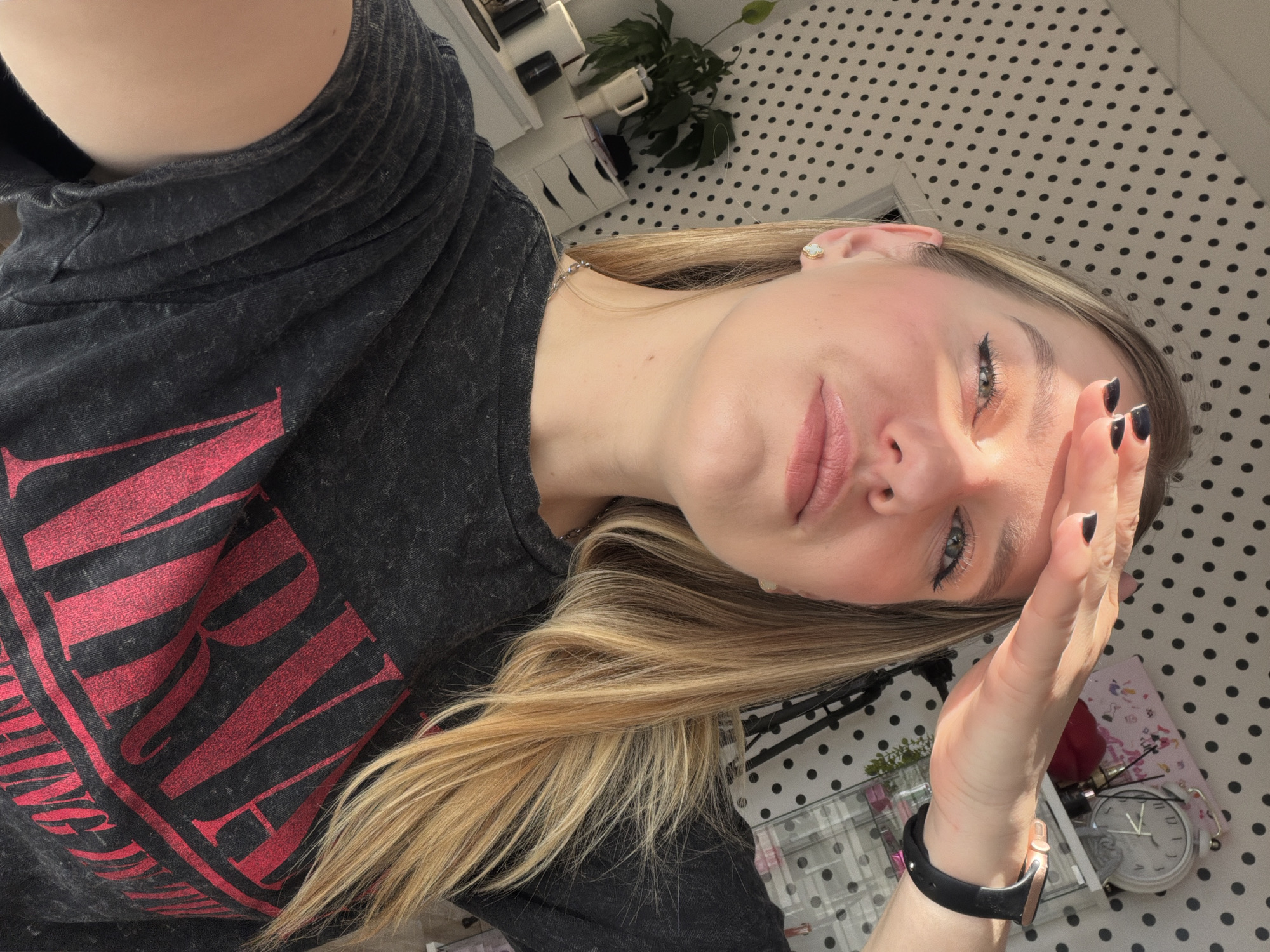 A selfie-style close-up shot, angled diagonally, captures a young woman shielding her eyes from bright sunlight. Her fair skin, light blonde hair, and blue-green eyes are prominent. She has a subtle make-up look with winged eyeliner and a soft pink lip color. She is wearing a dark gray, faded t-shirt with large red lettering, which partially reads "NIRVAN" (likely part of "NIRVANA"). Her left hand is raised, palm facing out, blocking some of the direct sunlight from her face. She has black nail polish on her short nails and a black-strapped smartwatch with a rose gold bezel on her left wrist. A small, light-colored flower-shaped earring is visible on her right ear.
The background suggests an indoor setting, likely a room in Riga, Latvia. Behind her, a surface covered in a light-colored fabric or wallpaper with black polka dots is visible. There are glimpses of white furniture with drawers, a small green potted plant, and other personal items such as a white analog clock, a pink notebook, and a glass organizer. The strong sunlight indicates it is daytime. The overall mood is casual and natural, with the direct sunlight creating bright highlights and soft shadows on her face.