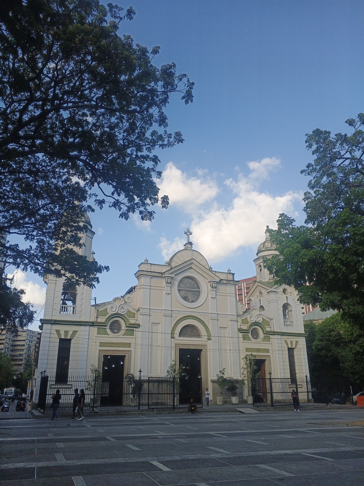 The image shows the Santa Teresa Basilica in Caracas, Venezuela. The Basilica is a large, ornate building with two bell towers, a central facade with a large round window, and a cross at the top. The building is painted in off-white with green trim. A black metal fence runs along the front of the building.
In front of the Basilica is a pedestrian area, with people walking and standing. Some people are near the fence. A tree with dense foliage partially obscures the left side of the Basilica. Another tree is on the right side of the building. The sky is blue with scattered white clouds. Other buildings are visible in the background.