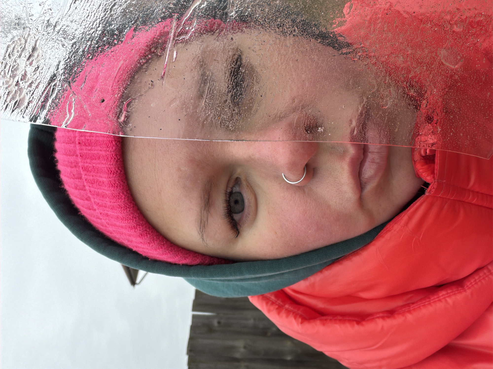 A close-up, rotated image shows a person wearing a bright pink knit headband and a red puffy jacket. Part of the person's face is obscured by a clear, textured sheet, possibly of ice or plastic. A silver ring is visible in the person's nostril. The background consists of a gray, overcast sky and a wooden fence. The location is Chaikovsky, Russia.