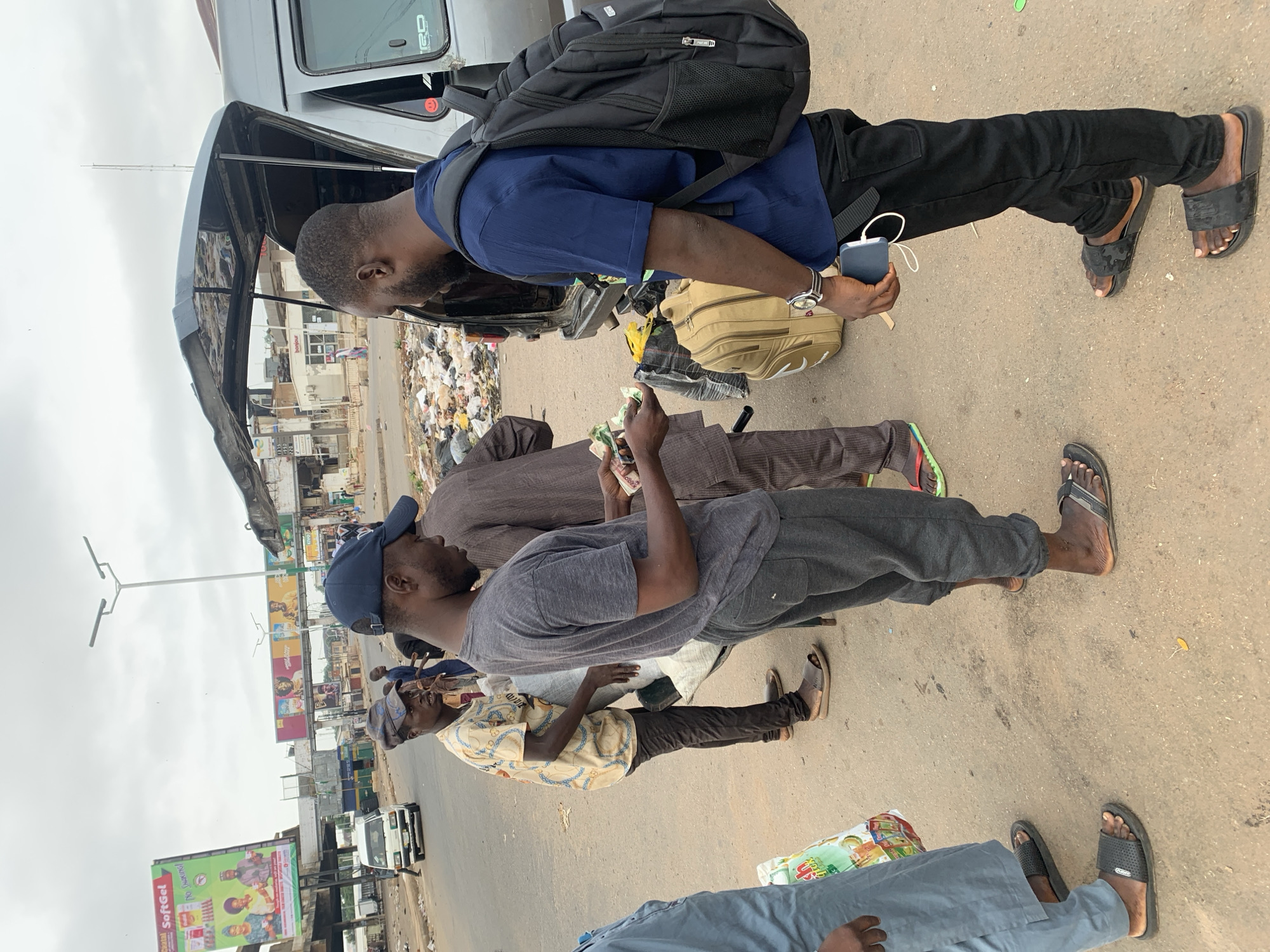 Jos, Nigeria: A group of men are gathered at a roadside bus stop. The bus in the image is loading passengers. The bus has trash behind it. The men are wearing casual clothes and sandals. Two of the men are holding money. One man is holding a mobile phone and has a backpack on his back. There are billboards in the background.