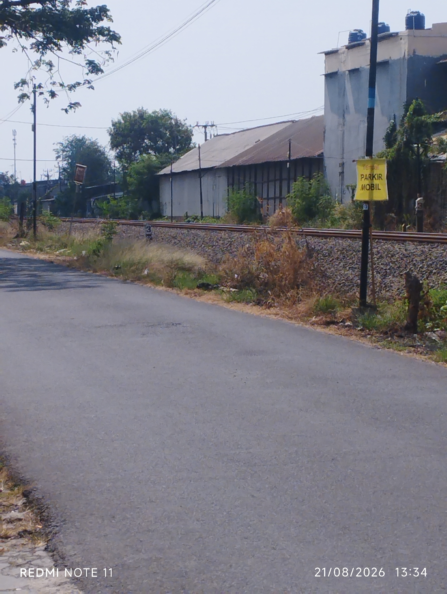 The image captures a street scene in Karawang, Indonesia, with a focus on a paved road running alongside a railway track. A yellow sign with Indonesian text, "TERTOP PARIK MOBIL," is prominently displayed on a pole, indicating a "Car Parking Area." The environment appears semi-industrial, with warehouses or large buildings visible in the background. Vegetation, including trees and dry grass, lines the railway tracks and the side of the road. The sky is clear and bright, suggesting daytime. A water tower is visible on top of one of the buildings. The bottom left corner of the image displays the text "REDMI NOTE 11," and the bottom right corner shows the date "21/08/2026" and time "13:34." There are no people or active vehicles visible in the frame.