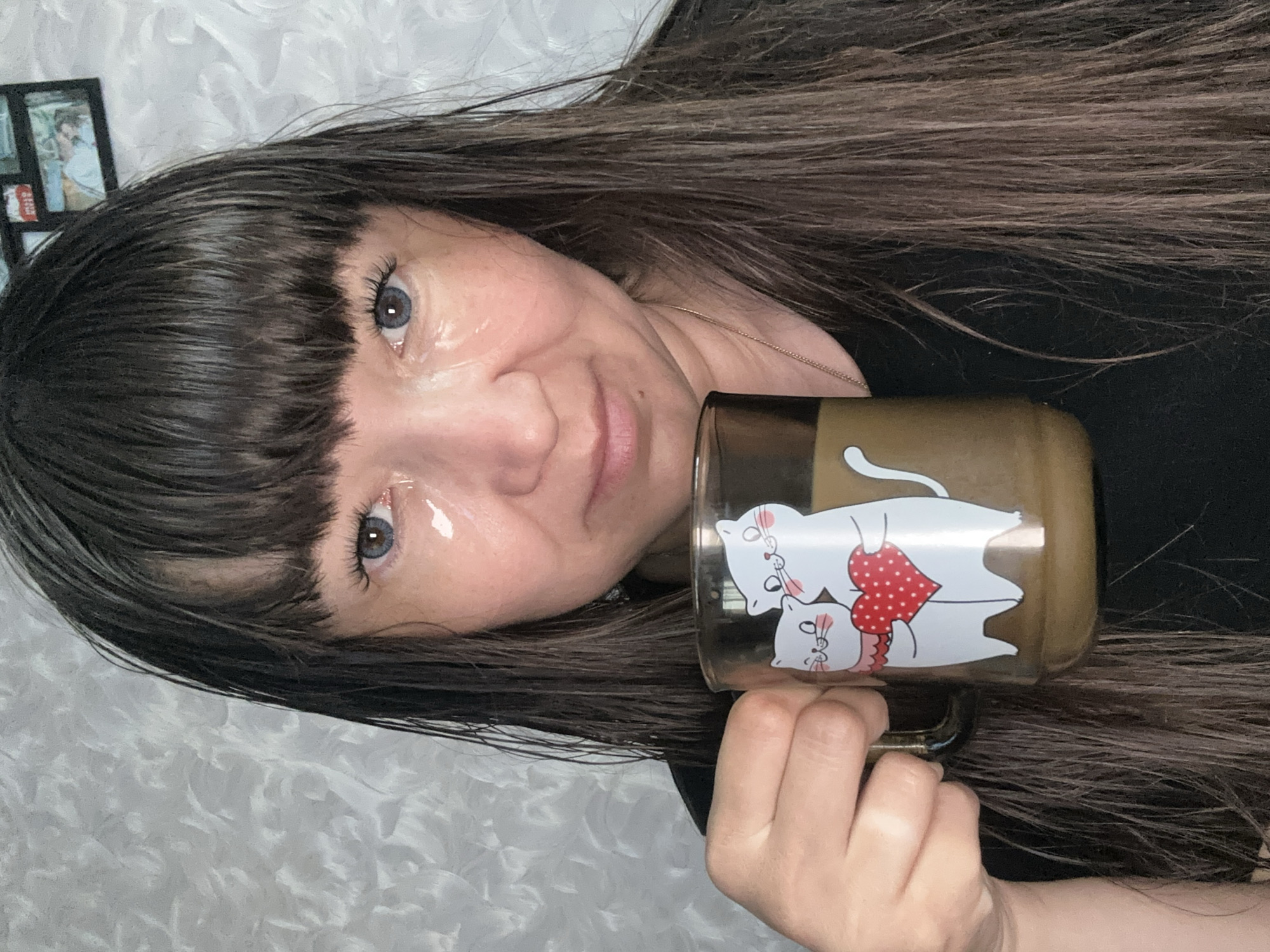 A woman with long dark hair and bangs holds a mug with a cat design. Her eyes are blue. The mug appears to be filled with a brown liquid, possibly coffee or tea. A small framed picture is visible in the background. The background is a textured, possibly white, material. There is no information given in the image that indicates its location as the city of Chaikovsky, Russia.