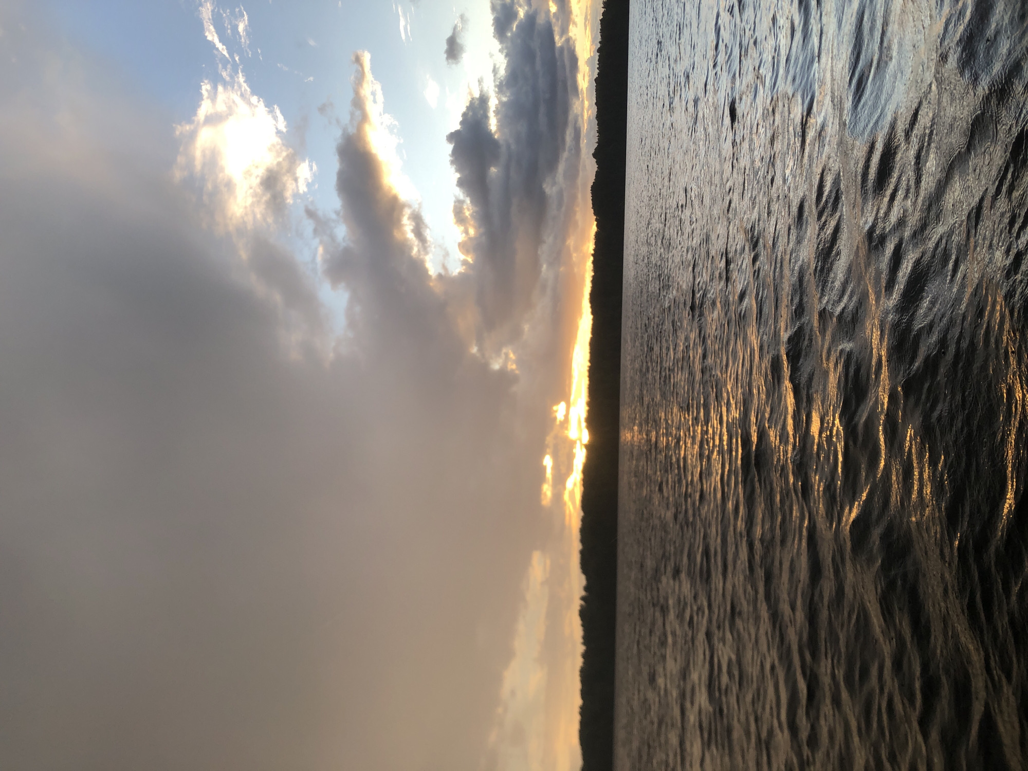 The image depicts a sunset over a body of water, likely a lake or a wide river, on the outskirts of Svirstroy, Russia. A dense, dark green treeline forms the horizon, separating the water from the sky. The sky is filled with dramatic clouds, with patches of bright orange and yellow sunlight breaking through, illuminating the water's surface with a shimmering reflection. The water itself is choppy, with small waves creating ripples and highlights. The overall impression is one of natural beauty and tranquility, characteristic of a rural or natural setting near Svirstroy. There are no people or man-made structures visible in the frame.
