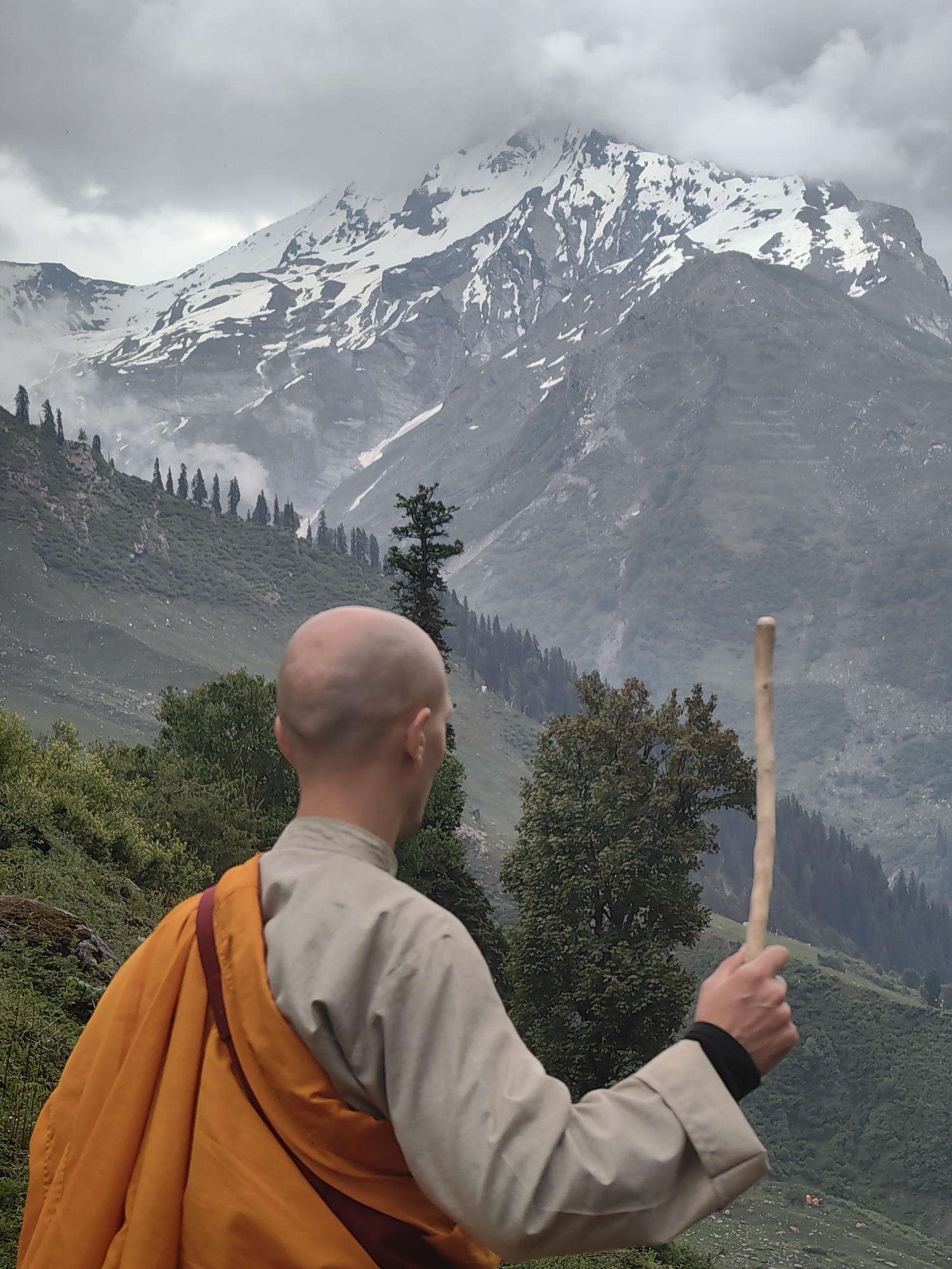 This media file depicts a lone individual in the foreground, viewed from behind, standing amidst a mountainous landscape. The subject has a shaved head and wears a light-colored robe draped with a vibrant orange cloth, possibly a monastic garment. They are holding a simple wooden stick in their right hand.
The background is dominated by a majestic, snow-capped mountain range under a cloudy sky. The slopes of the mountains are covered in lush green vegetation and scattered trees, with patches of snow visible on the higher elevations. Wisps of mist or fog add a dramatic and atmospheric quality to the scene, particularly in the mid-ground and around the mountain peaks. The overall setting suggests a remote, high-altitude location, consistent with the provided location context of Dhundi, India.
The weather appears overcast, with a diffused light suggesting it could be morning or late afternoon. The mood conveyed is one of solitude, contemplation, and awe inspired by the grandeur of nature. The activity taking place is minimal, focusing on the individual's presence within the vast natural environment, perhaps on a pilgrimage or a journey. There is no visible text in the image.