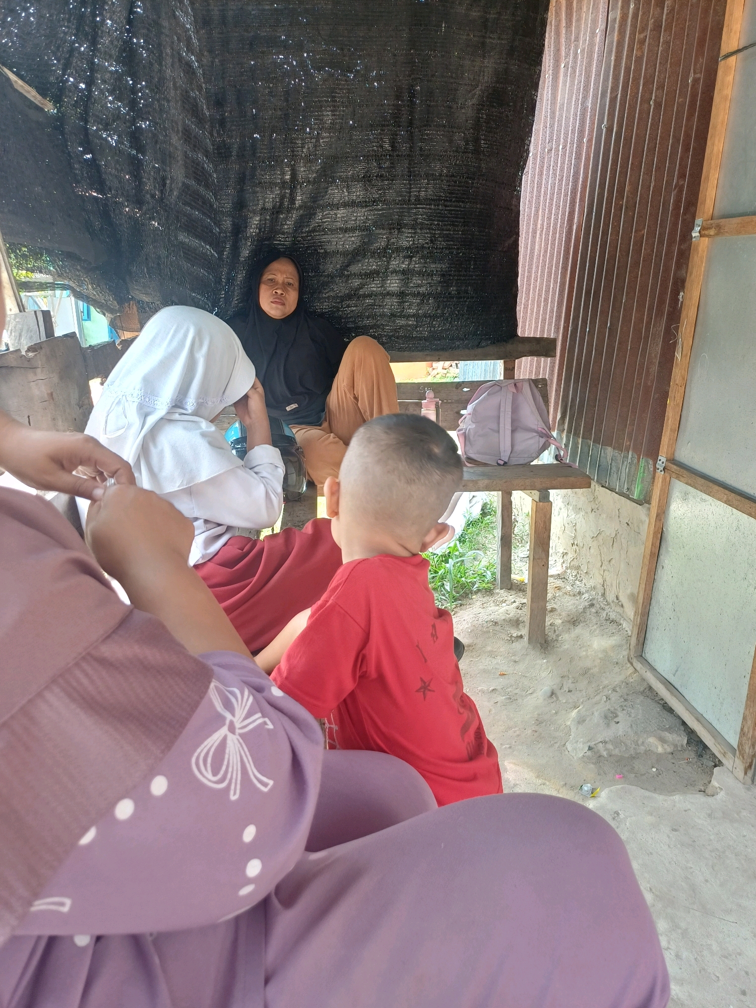 In Dumai City, Indonesia, an outdoor scene unfolds under a makeshift canopy of black netting. Three individuals are present, including a woman in a black hijab and brown pants seated on a wooden bench, her gaze directed forward. To her left, a child wearing a white hijab and red skirt sits, seemingly engaged in adjusting their headwear. In the foreground, a young boy in a red t-shirt with a star print is seen from behind, facing away from the viewer and towards a dusty ground surface. A light pink backpack rests on the bench beside the seated woman. The environment is rustic, with corrugated metal siding and rough-hewn wooden structures forming part of the background. The ground is composed of dirt and sand, with sparse vegetation visible.