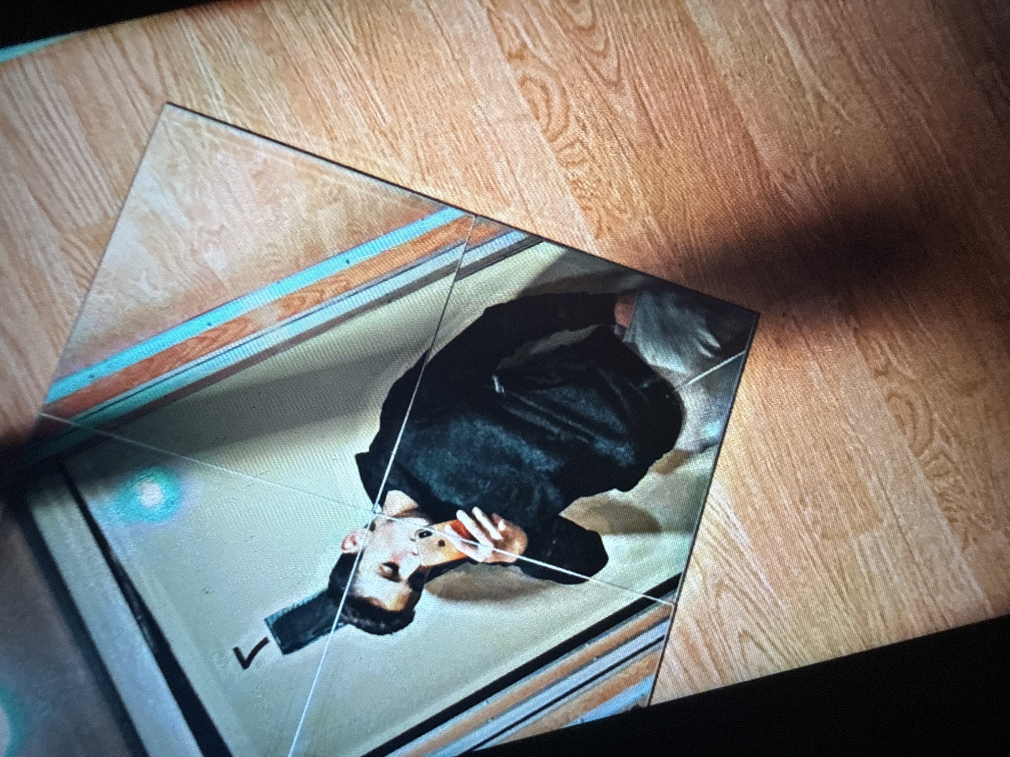 A man is taking a selfie in what appears to be a reflective surface, possibly a mirror in a room with wood-paneled walls. He is wearing a dark hooded sweatshirt and light-colored pants, holding a mobile phone in his right hand. The phone's screen is illuminated, and he is looking down at it. There is a noticeable crack in the reflective surface, running diagonally across the image. The background is indistinct, with some blurred shapes and colors that might indicate lighting or other objects in the environment. The floor visible in the foreground has a wood grain pattern. The image does not contain any discernible landmarks or clues that would help identify a specific location in Sofia, Bulgaria.