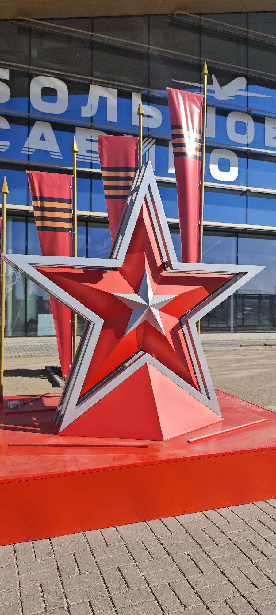 A bright red star monument sits in front of a building in Bolshoye Savino, Russia. The star is outlined in gray and sits on a red platform. Behind the star are three red flags with orange and black stripes. The building in the background has blue and white lettering that reads "БОЛЬШОЕ САВИНО". The ground is paved with gray brick.
