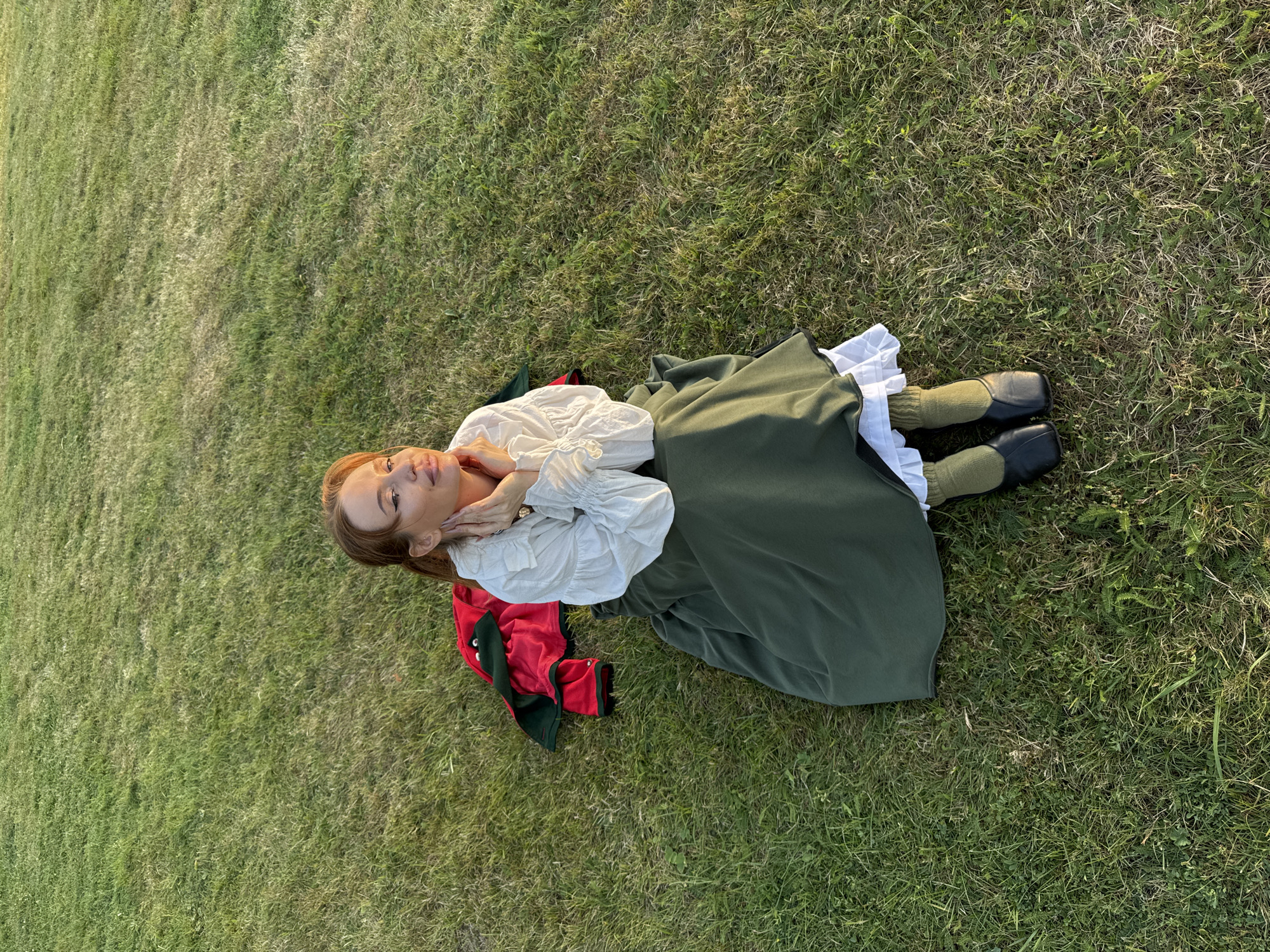 A woman with reddish-brown hair is sitting on a grassy field at the Estate Museum of V.I. Arkhangelskoye, Moscow Oblast, Russia. She is wearing a long, dark green skirt, a white ruffled blouse with puffed sleeves, green socks, and black shoes. A red and green jacket is laid on the grass next to her. The woman has her hands clasped under her chin and is looking upwards, away from the camera. The sun is casting a warm glow on the scene.