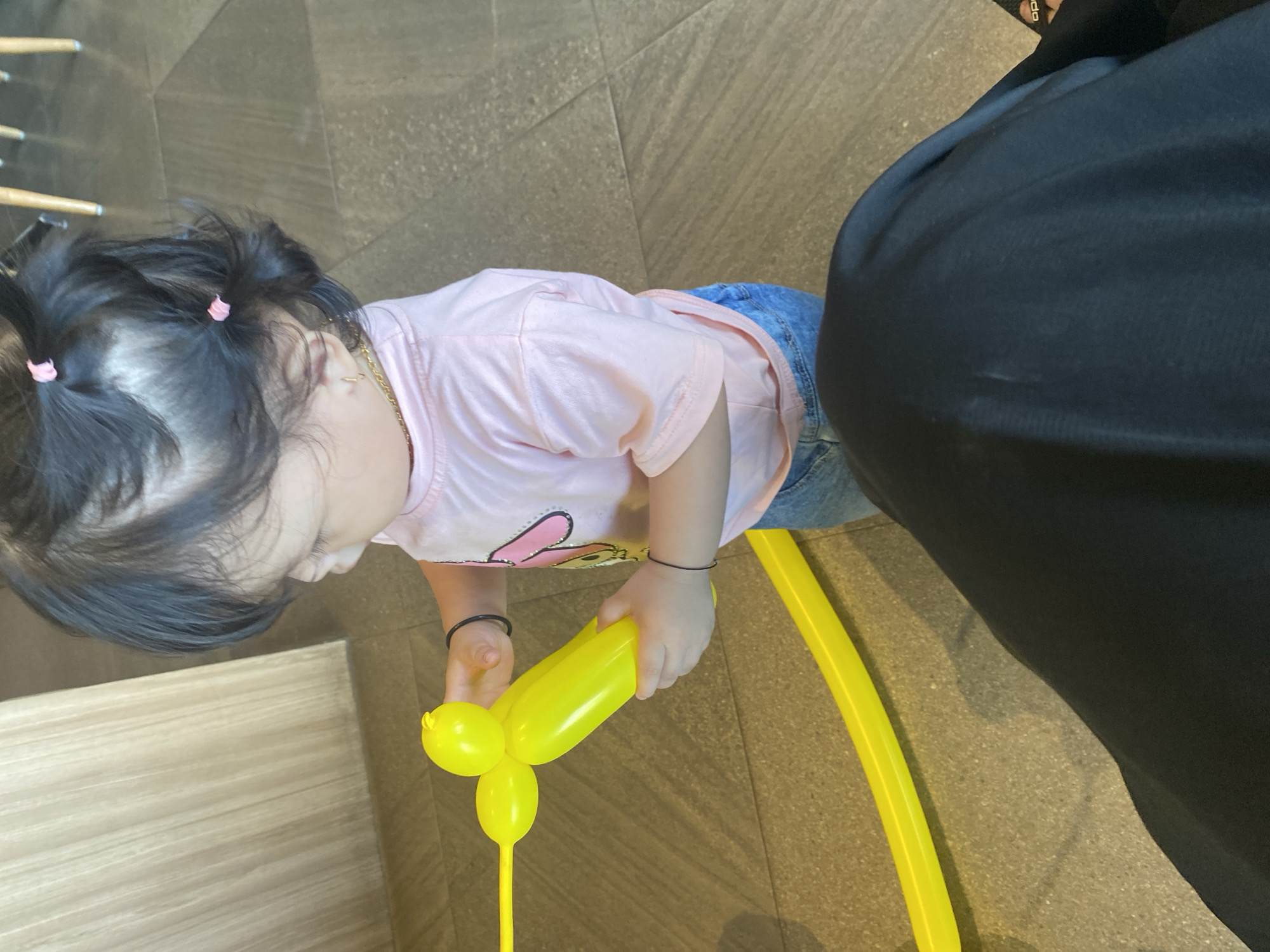A toddler, possibly female, with dark hair styled in pigtails with pink ties, stands and holds a yellow balloon animal. The child is wearing a light pink t-shirt and blue jeans. The background consists of a tiled floor and a light-colored wooden wall. The image appears to be taken indoors, likely in a public space. The toddler is looking down at the balloon animal. The scene is captured in Watampone, Indonesia.