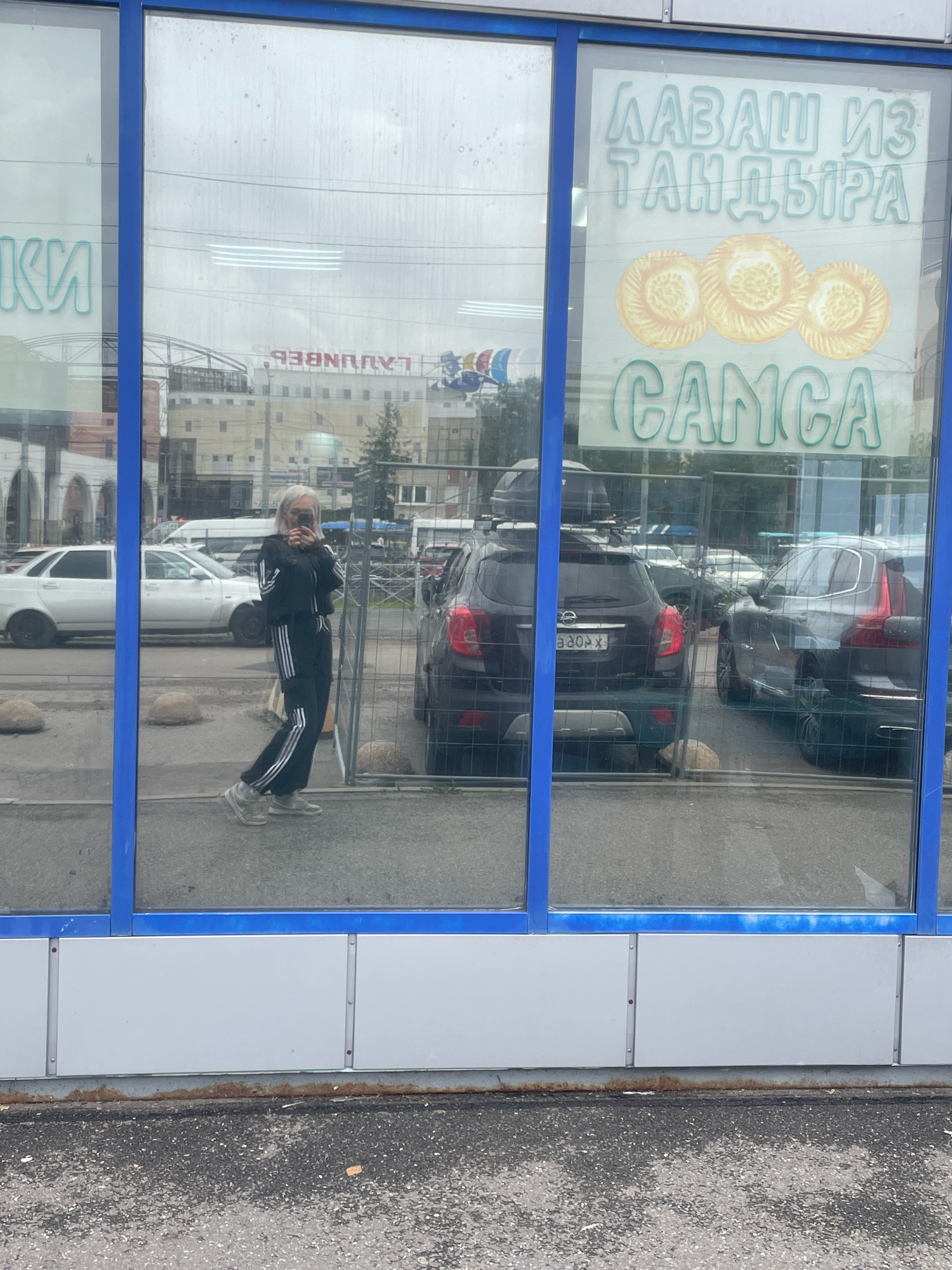 In Saint Petersburg, Russia, a young woman with blonde hair, wearing a black tracksuit with white stripes and grey sneakers, takes a selfie in a shop window. The window displays a sign with blue text that reads "ЛАВАШ ИЗ ТАНДЫРА" and images of three golden flatbreads, followed by "СAMCA" in green text. Behind her, the reflection shows cars parked on a street, including a black Opel, a grey Volvo, and a white car. The shop front features large glass windows framed by blue metal. The surrounding environment appears to be urban, with buildings and possibly a street visible in the reflection.