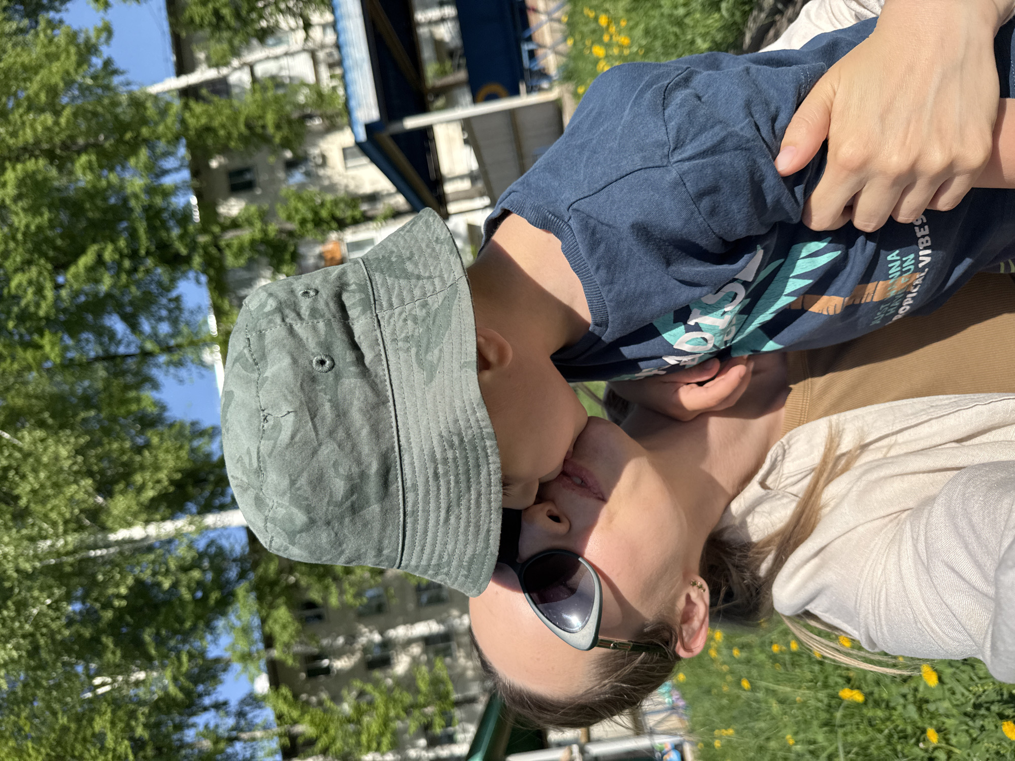 The image shows a person, likely a parent, wearing sunglasses and a light-colored shirt, embracing and kissing a young child with a bucket hat and blue t-shirt that reads "tropical vibes." The location appears to be outdoors on a sunny day, with green grass and yellow wildflowers visible. In the background are trees, a building, and a play structure. Based on provided information the location is the city of Chaikovsky, Russia.