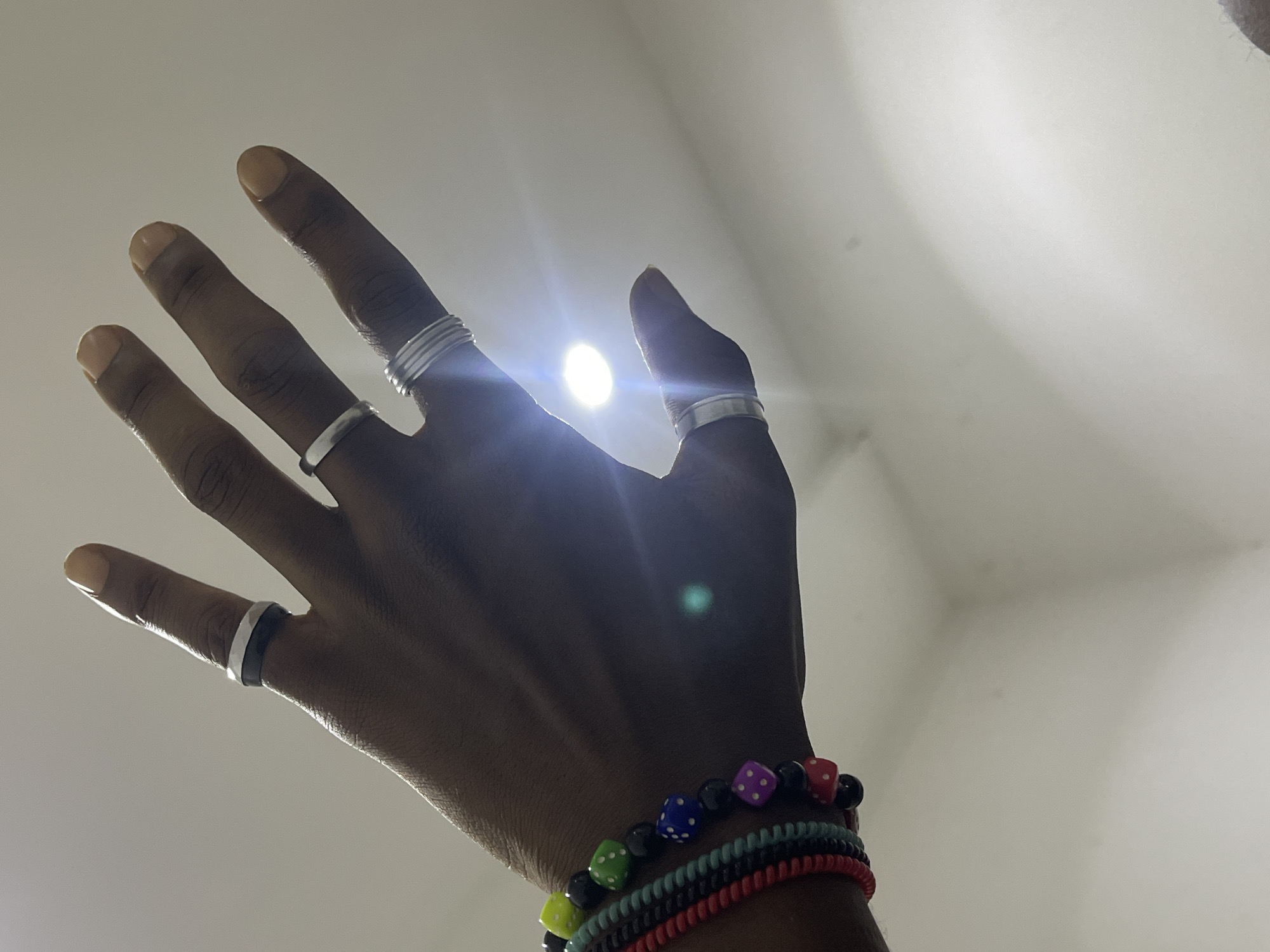 In Abuja, Nigeria, a close-up shot captures a person's left hand, adorned with several rings and bracelets, against a softly lit, off-white background. The hand is positioned with fingers extended and slightly spread. On the index finger, a silver ring with a black insert is visible. The middle finger sports a plain silver band, and the ring finger is encircled by a stacked set of three thin silver bands. The thumb is partially obscured, but a silver ring is noticeable on it. Around the wrist, a collection of colorful beaded bracelets is worn, including one with black beads and small, multi-colored dice-shaped charms, as well as other bracelets in red and teal. A bright light source causes a lens flare and a distinct glint on the back of the hand. The overall impression is one of personal adornment.