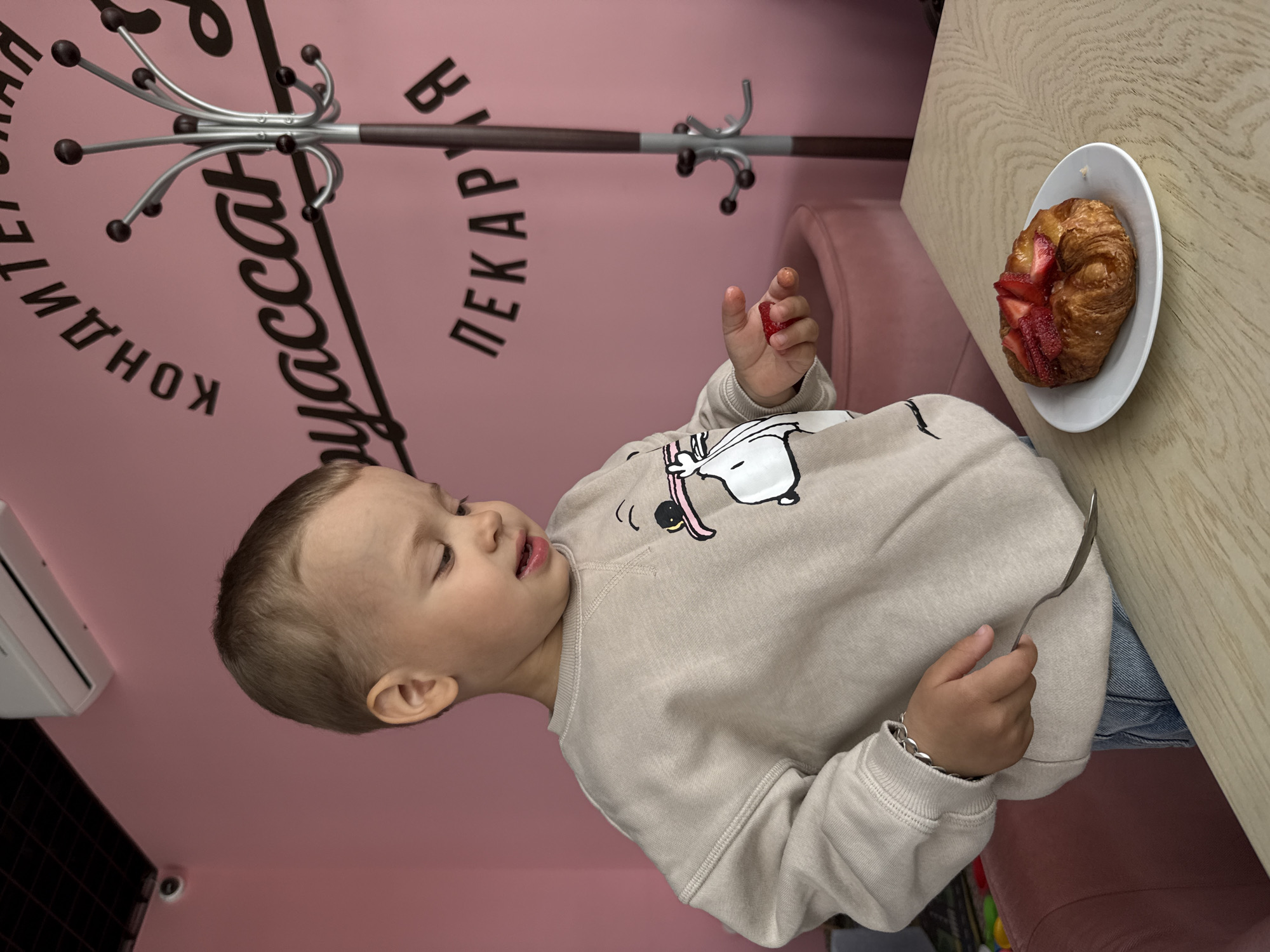 A young child with fair skin and short blond hair is seated at a light wood-grained table in what appears to be a café or restaurant. The child is wearing a light beige sweatshirt with a white cartoon graphic. In one hand, the child holds a small piece of red fruit, and in the other, a metal spoon. A croissant topped with sliced strawberries rests on a white plate on the table. The wall in the background is pink and features black cursive lettering in Russian: "Кондитерская пекарня" (Confectionery Bakery). A coat rack is attached to the wall above the child. An air conditioning unit is visible on the wall to the child's left. This scene is likely in Rusanovo, Russia, given the Russian inscription on the wall.