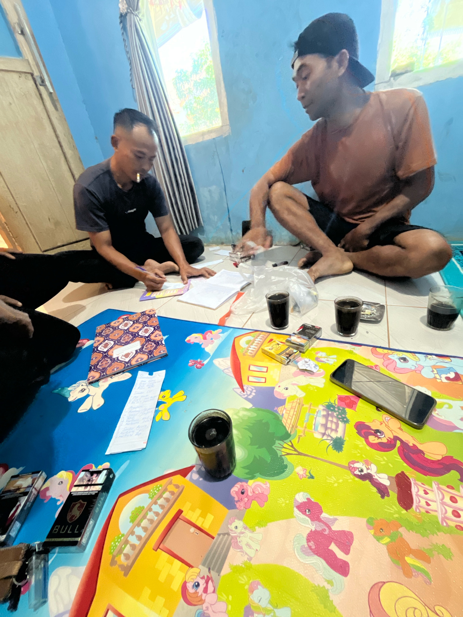 Two men are sitting on the floor in a room with blue walls. The man on the left is wearing a dark t-shirt and has a cigarette in his mouth. He is writing on a piece of paper with a pen. The man on the right is wearing an orange t-shirt and a black baseball cap. He is looking down at his hands. In front of them is a colorful mat with images of cartoon ponies. On the mat are several glasses of dark liquid, packs of cigarettes, a lighter, and a smartphone. The room appears to be in a residential setting in Margodadi, Indonesia.