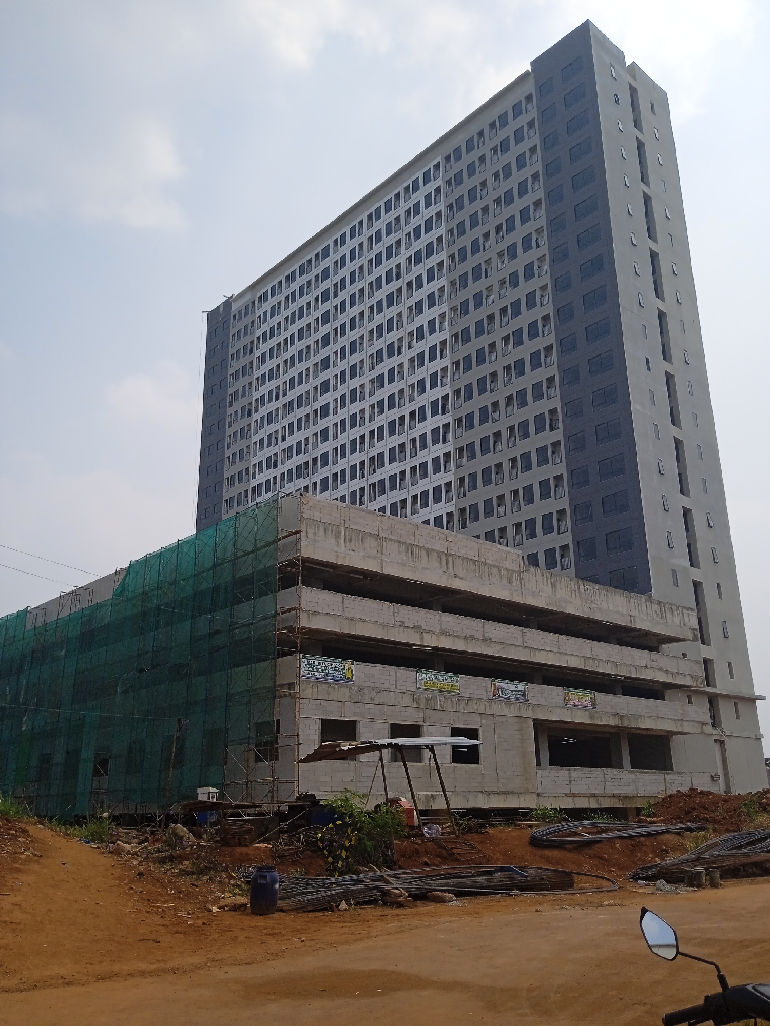 A tall, modern skyscraper, under construction, dominates the frame, its facade a grid of numerous dark-framed windows. The building appears to be in a partially completed state, with scaffolding and green netting visible on lower levels, suggesting ongoing construction. Adjacent to the skyscraper, a multi-story structure is also under construction, with visible concrete floors and brick walls. Several banners with text are attached to this lower building. The surrounding area is a dusty construction site with piles of dirt, construction materials like rebar, and a blue plastic barrel. The sky is overcast with scattered clouds. The scene depicts an urban development project in progress in Bekasi, Indonesia.