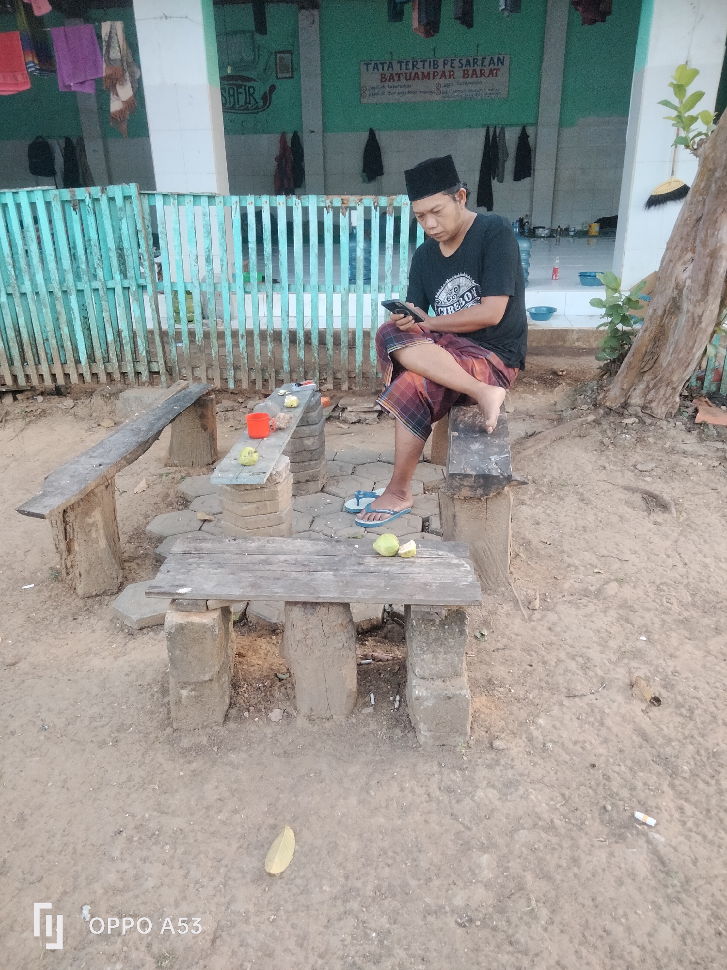 The image depicts an outdoor scene in what appears to be a village setting in Badung, Indonesia. In the foreground, a man wearing a black t-shirt, sarong, and peci (a traditional cap) is seated on a wooden bench, looking at his mobile phone. In front of him, a rustic table made of wood and concrete blocks holds an orange cup and two guavas, one of which is sliced. Another similar wooden bench is situated to the left, and a lower bench is in the immediate foreground. The background reveals a building with green walls and a white pillar. A wooden fence with vertical turquoise slats runs across the middle ground. Various items are hanging from the building's exterior, including clothes and a broom leaning against a tree. The ground is covered in dirt and small stones, with a few cigarette butts visible. A water bottle and a blue bowl are also present near the building entrance. The overall environment suggests a casual, possibly religious or communal gathering space.