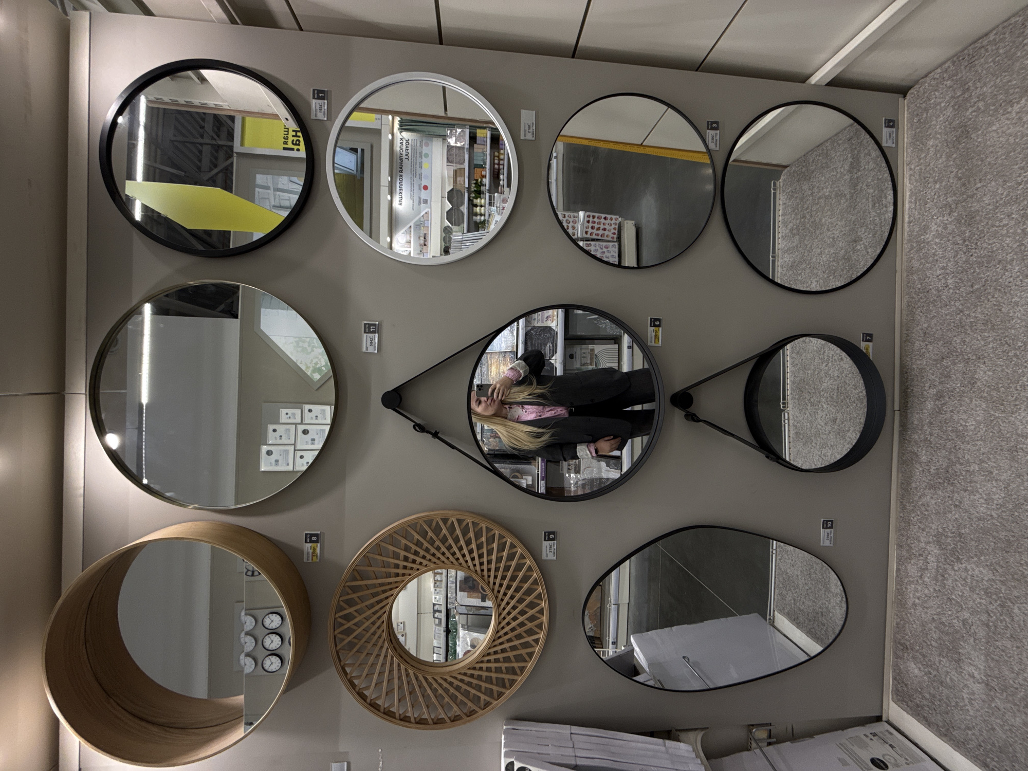 The image shows a display of nine different mirrors on a gray wall, likely inside a store. The mirrors vary in shape, size, and style, including round, oval, and decorative frames. A person is visible in the reflection of one of the mirrors, holding a phone, presumably taking the photograph. The reflections in the mirrors show the surrounding store environment, including shelving and other products. The presence of price tags and product information sheets adjacent to some of the mirrors suggests that they are for sale. The overall setting seems to be a retail environment showcasing home decor items. I don't see details that would indicate this scene is in Izhevsk, Russia.