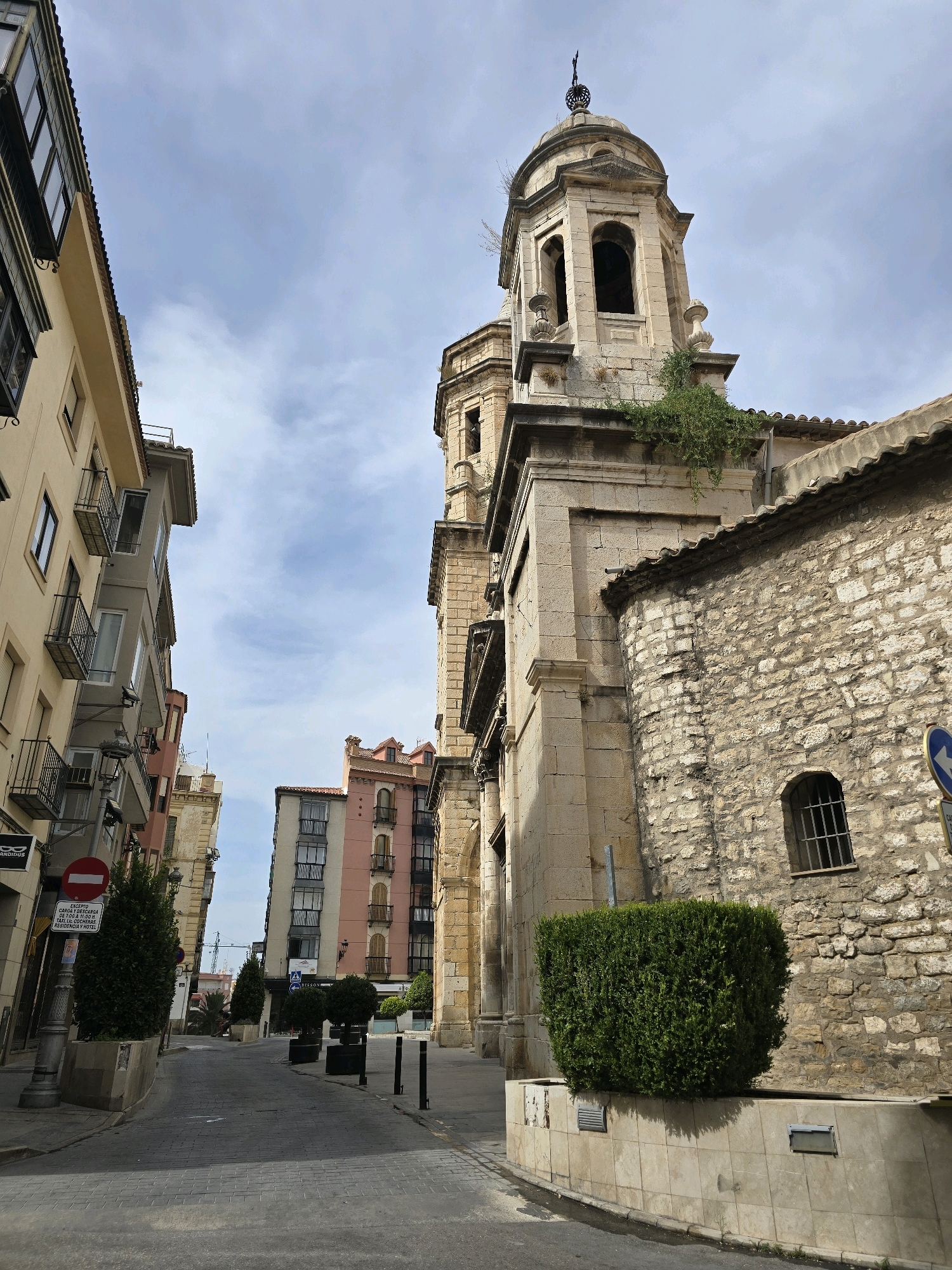 An eye-level, outdoor, vertical, full shot captures a narrow cobblestone street in an urban European setting under a partly cloudy sky. The street is lined with buildings on both sides, with a prominent stone church featuring a bell tower dominating the right side of the frame. The church's facade is constructed from rough-hewn stone blocks, with a tall, arched window and a tiled roof. Lush green foliage grows from the upper levels of the church. On the left, a building with a cream-colored facade features multiple balconies with black wrought-iron railings and dark-framed windows. Further down the street, a building with a pink and cream facade presents a similar architectural style.
The street itself is paved with cobblestones and is devoid of people. Planters containing neatly trimmed evergreen bushes are strategically placed along the sidewalk. A street lamp with a sign is visible on the left, and a blue and white directional sign is partially visible on the right. The overall impression is one of a historic European city with a blend of residential and religious architecture.