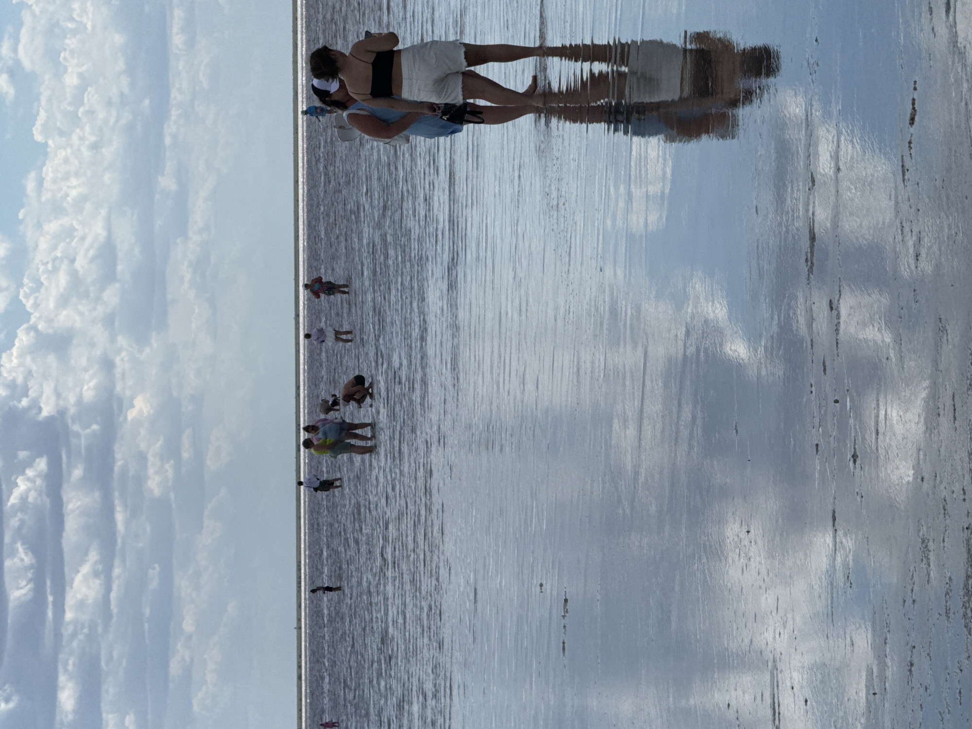 In a shallow, reflective body of water, several people are wading and interacting. In the foreground, two women are standing close together, one with a white baseball cap and black tank top, the other in a blue dress, both with their backs to the camera. Their reflections are visible in the water. Further back, a group of people are scattered across the water. One man in a white shirt and dark pants stands near the center. To his left, a group of three people in casual clothing are wading, one of whom appears to be crouching. More individuals are visible in the distance, including a person in pink and another in blue. The water is calm and mirrors the cloudy sky above, which is filled with large, white cumulus clouds. The horizon line is clearly defined, separating the water from the sky. The environment appears to be a natural body of water, possibly a salt lake or lagoon, with a flat, sandy or muddy bottom visible in places. The location is identified as Bursol, Russia.