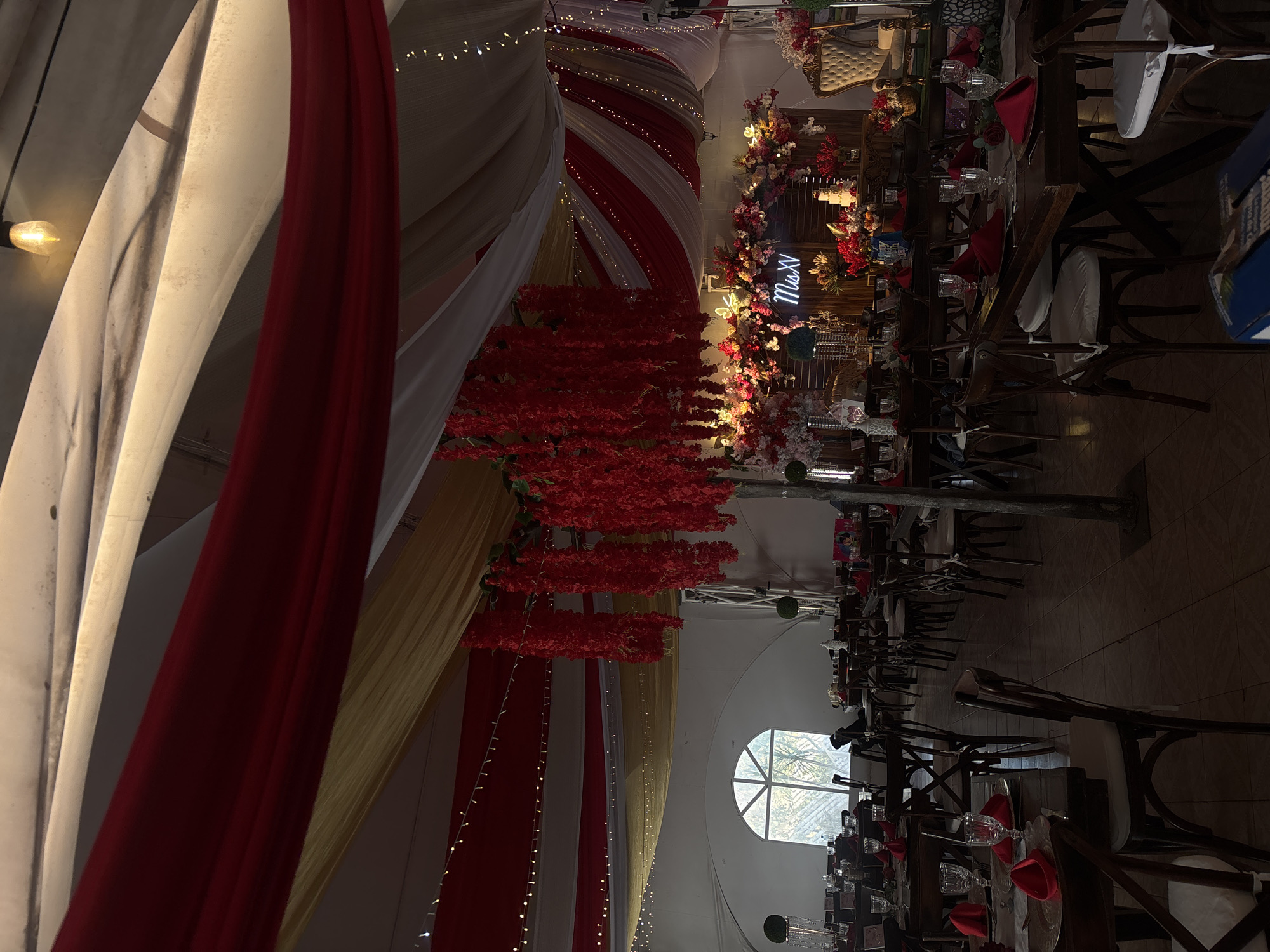The image depicts an interior event space in Chilpancingo, Mexico, likely a quinceañera celebration. The room is decorated with elaborate draping in red, white, and gold fabrics, accented with string lights and hanging red floral arrangements. Tables are set for a formal meal with glassware, red napkins, and dark wooden chairs. The overall lighting is dim. A window in the background provides natural light. The focal point appears to be a stage or dais further into the room, decorated with flowers and a neon sign. The room is set up for a celebration.