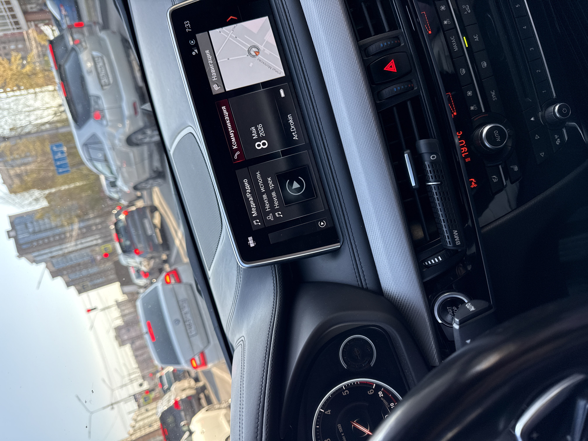 The image shows the interior of a BMW car, focusing on the dashboard and infotainment system. The infotainment screen displays a navigation map indicating the location is near "улица Молокова" (Ulitsa Molokova or Molokov Street). It shows a date in the future (May 8, 2026) and the name "Art.Drokin." The car's climate control panel is visible, displaying a temperature of 19.0°C. The car is in traffic, as seen through the windshield, with other vehicles and buildings visible outside. A small air freshener is clipped to one of the vents.