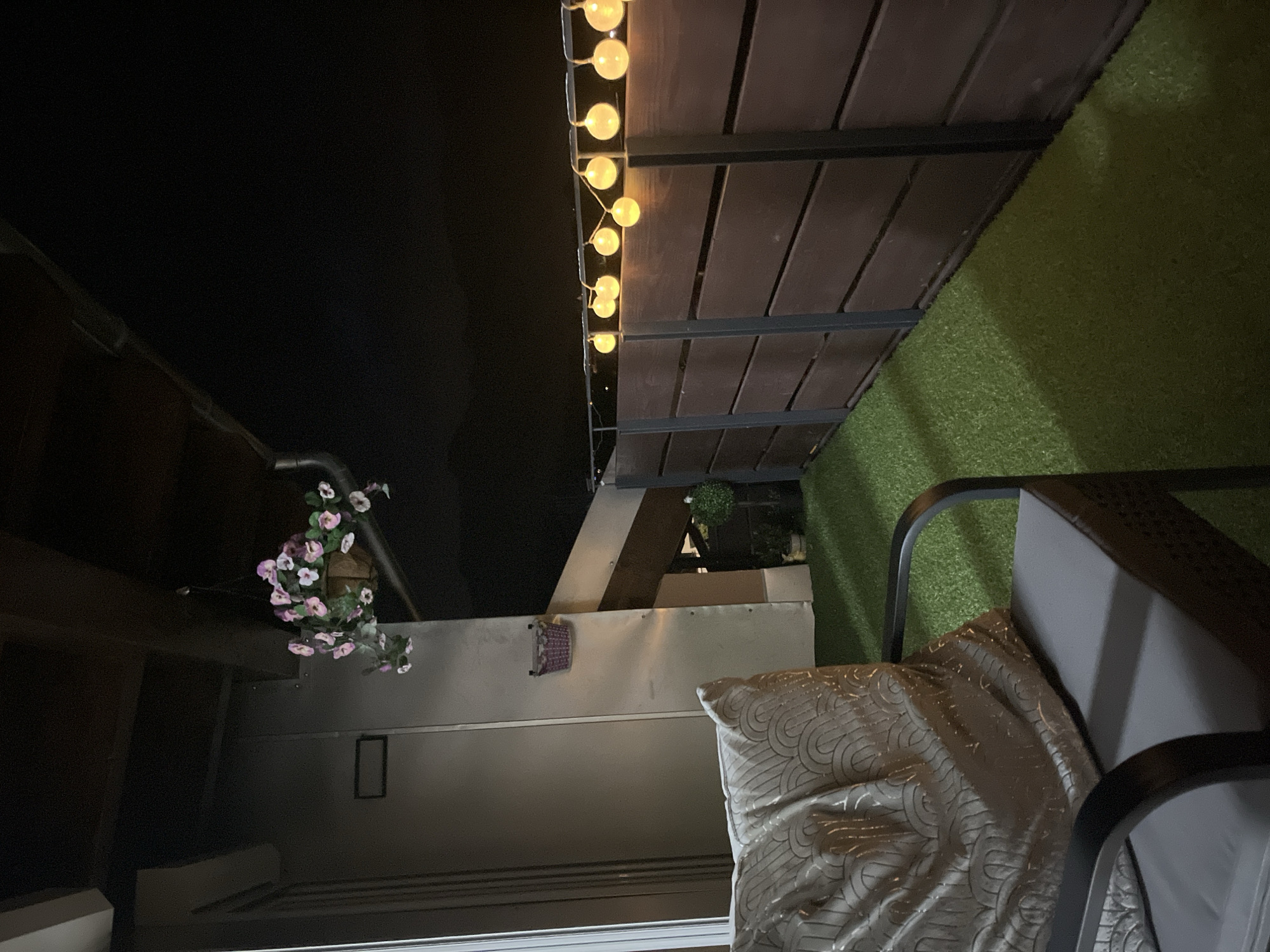 This image depicts a balcony or patio area at night. Artificial turf covers the floor, and a dark wooden fence runs along the perimeter. String lights with round, yellow bulbs are hung along the top of the fence. In the foreground, a chair with a gray cushion is partially visible, topped with a decorative pillow featuring a geometric pattern. To the left, a metallic wall or structure is present. Above, a hanging basket filled with pink and white artificial flowers is suspended. A small, decorative purple basket is mounted on the metallic wall. In the background, a dark, out-of-focus landscape suggests the presence of mountains, consistent with the location in Garmisch-Partenkirchen, Germany. The lighting is dim, with the string lights providing the primary illumination. There are no people visible in the image.