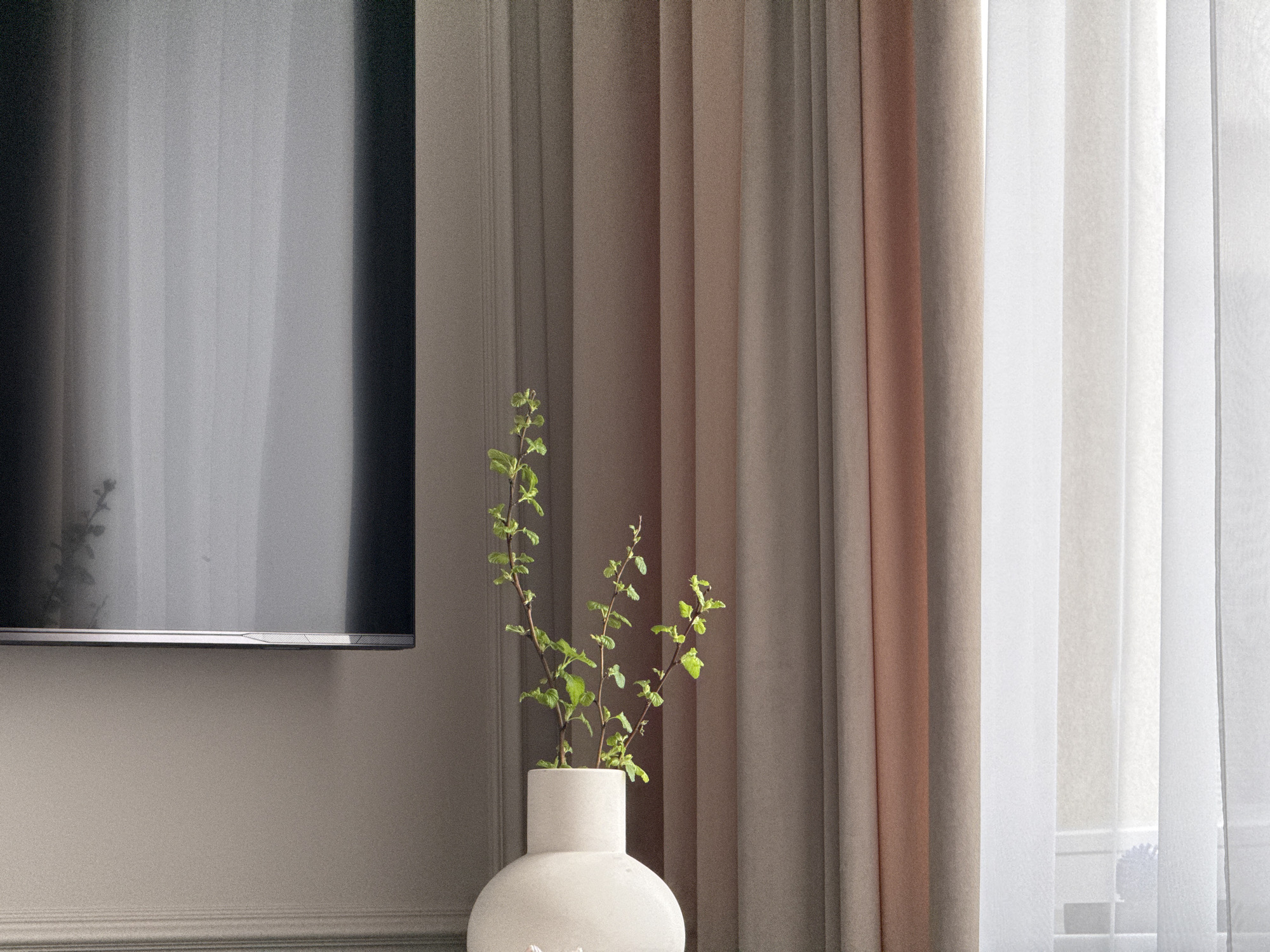 The image shows a room interior. A flat-screen television is mounted on a wall painted in a neutral tone. To the right of the television, curtains hang in varying shades of beige, light brown, and white, creating a layered effect. In the foreground, a white vase is visible, holding green leafy stems. The vase is placed on what appears to be a ledge or surface, and the stems reach upward, partially obscuring the view of the curtains behind. The lighting in the room is soft and diffused, lending a calm and serene atmosphere to the scene.