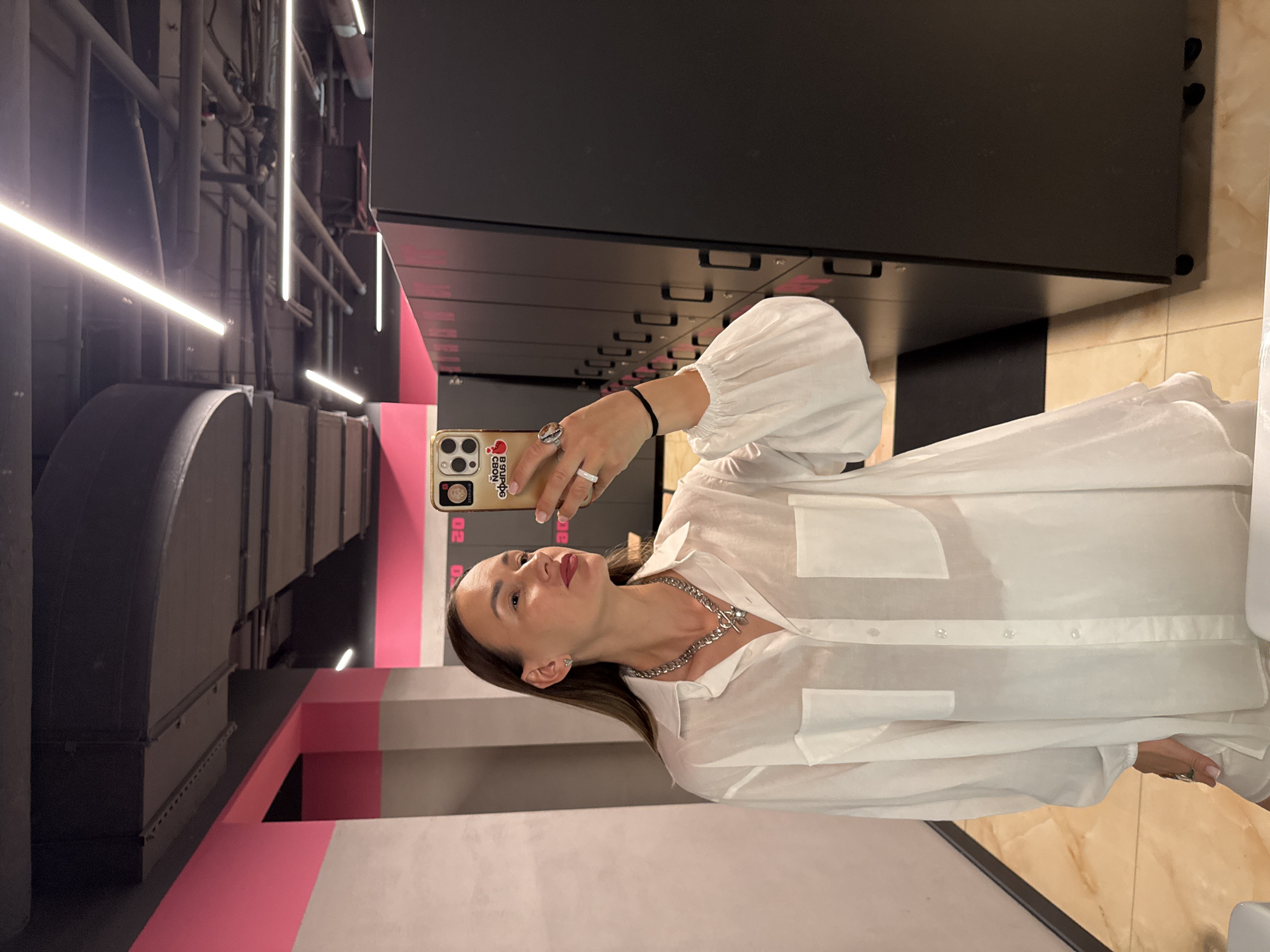 A woman takes a selfie in a locker room, holding her phone in her left hand. She is wearing a white button-up shirt with large pockets and puffy sleeves. She has on a chunky silver necklace and rings on her fingers. Her hair is brown and falls past her shoulders. The locker room has black lockers with silver handles and pink accents. There are pipes and lights visible on the ceiling. The floor is tiled with a marble-like pattern. The woman has a slight smile on her face and appears to be in good spirits. The image seems to have been taken indoors, likely in a gym or spa. There is some text visible on the phone case, but it is too blurry to read. The overall impression is a casual and confident pose in a modern locker room setting.