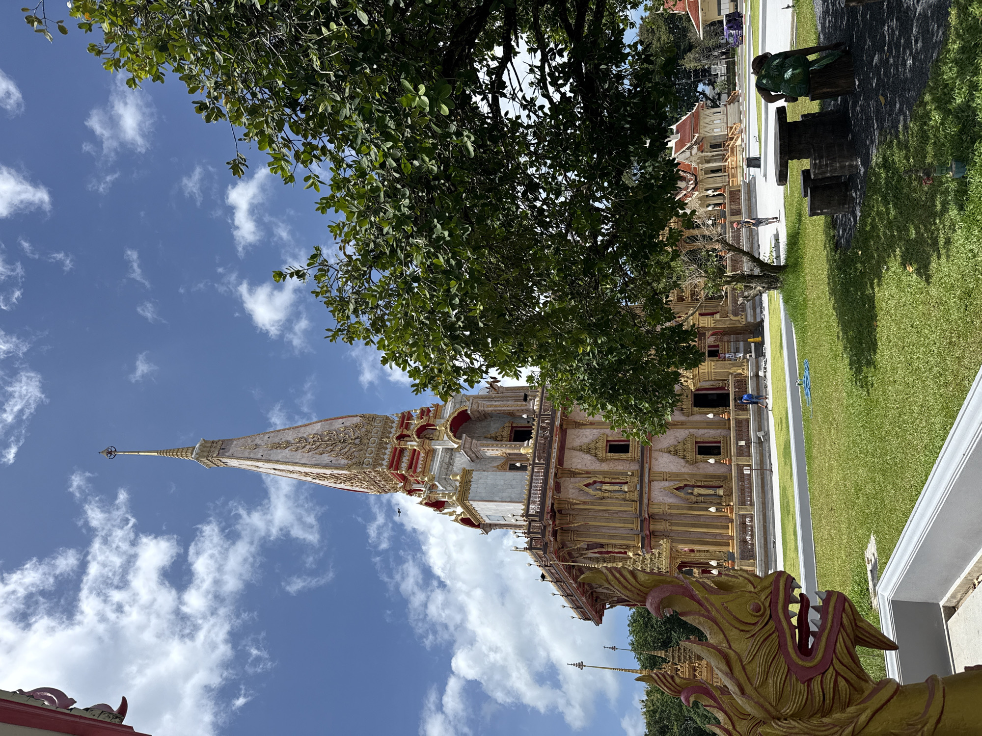 A full shot of a Buddhist temple, Wat Chalong, in Chalong, Thailand, is captured on a bright, sunny day with scattered clouds. The temple's prominent white and gold stupa rises majestically against the blue sky, adorned with intricate golden patterns and red accents. In the foreground, a vibrant green lawn is dotted with a few trees and a decorative statue of a mythical creature. To the right, a rustic outdoor seating area with a wooden table and tree stump seats is visible. In the middle ground, people can be seen walking around the temple grounds. The overall scene exudes a sense of tranquility and architectural splendor.