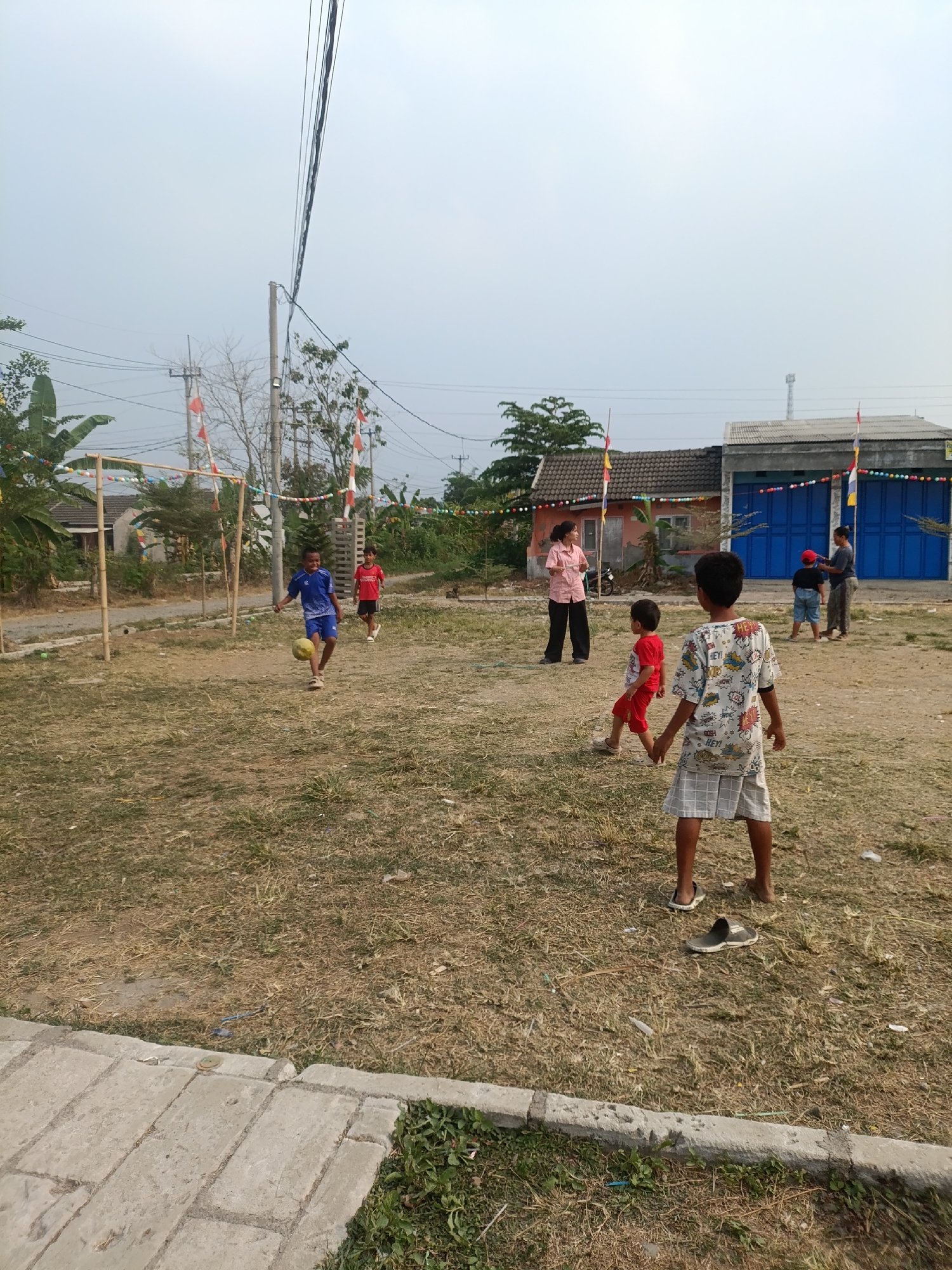 In Cariu, Indonesia, a group of people are gathered in an open, grassy area with dry vegetation. The scene appears to be a casual gathering or recreational event, possibly in a residential neighborhood given the presence of houses in the background. A makeshift soccer goal constructed from bamboo poles stands on the left side of the image. Several individuals, predominantly children, are engaged in playing soccer. One boy in a blue shirt and shorts is actively kicking a yellow soccer ball. Another boy in a red shirt and shorts is nearby. In the background, two adults and two other children are visible. One adult, wearing a pink shirt and dark pants, stands to the left, possibly observing the game or interacting with the children. To the right, another adult in darker clothing stands near a blue-doored building, and a child with a red hat is also present. The environment is characterized by an overcast sky, some trees and vegetation, and power lines running overhead. Decorative flags and colorful garlands are strung between poles, suggesting a festive occasion. The ground is predominantly dirt and dry grass, with some scattered debris. A concrete pathway runs along the bottom of the image, bordering the grassy area.