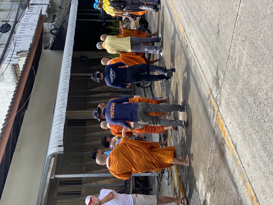 This forensic description details a daytime street scene in Ban Phe, Thailand. A procession moves along a concrete street with faded yellow lines and visible stains. Approximately four bald, barefoot Buddhist monks, draped in saffron/orange robes, are central to the image. They are engaged in interaction with civilians, consistent with an alms round.
On the far left, a light-skinned man in a white polo shirt, beige shorts, and sandals stands, observing the group with his left arm raised. Towards the center-left, behind the leading monk, civilians including a person in a patterned top are seen extending their hands towards the monks, suggesting alms giving.
Two men in dark blue long-sleeved shirts are prominent. One, in the center, wears grey trousers and a backward baseball cap, with "กรมประมง" (Department of Fisheries) in white Thai script on his back. Another man in a similar dark blue shirt with a white emblem and backward cap stands further right. These individuals appear to be officials or organizers.
Other participants include a man in a light yellow polo shirt and light grey trousers on the right, and several individuals in yellow shirts and blue caps further right, one with a black backpack.
The background consists of a row of older two-story buildings, likely shophouses, with closed ground-floor storefronts and a continuous corrugated metal awning extending above. Overhead, electrical wires are visible. A white scooter is partially visible on the right side of the street. The bright sunlight casts distinct shadows on the ground and subjects.