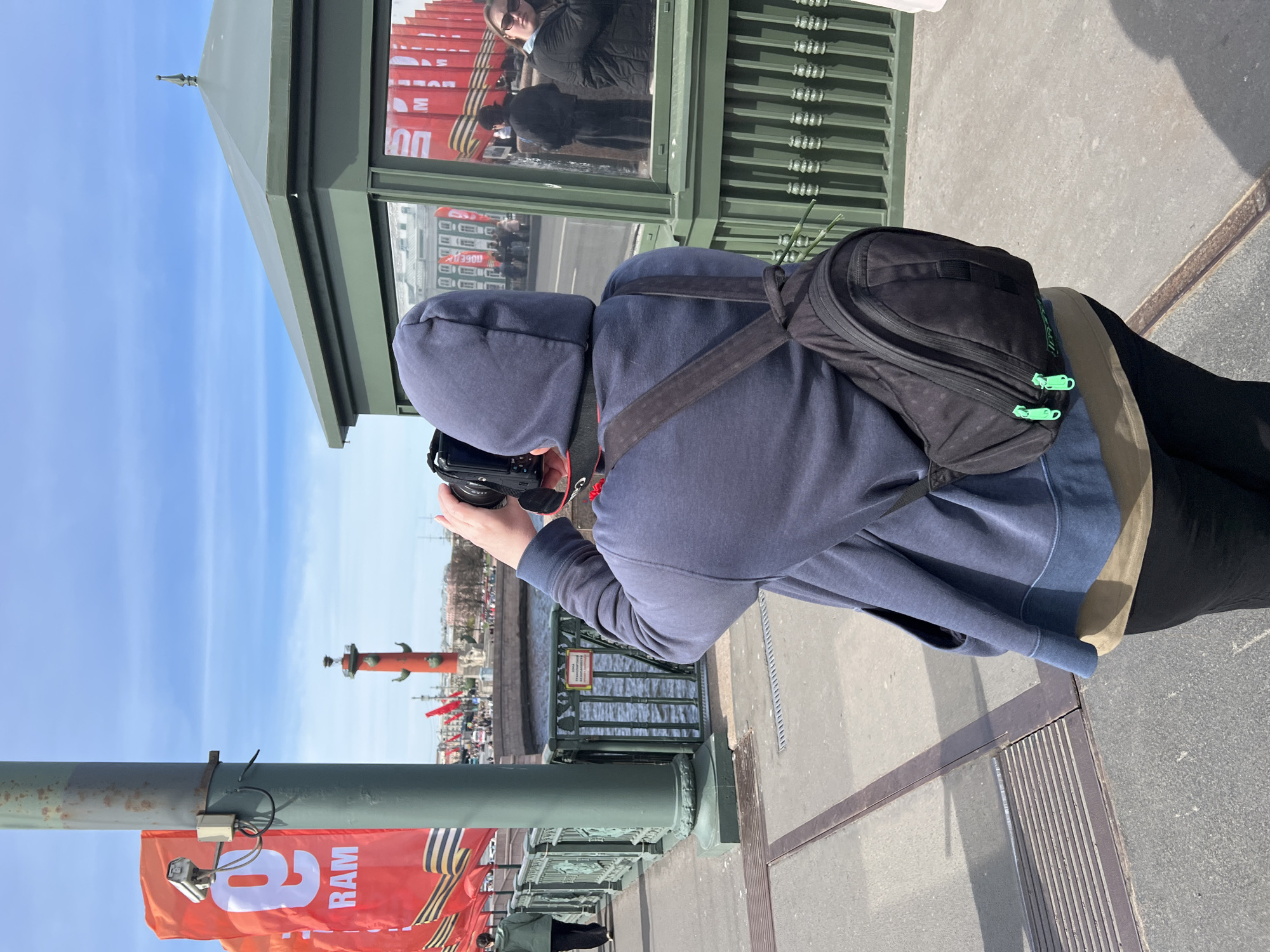 A person wearing a blue hooded sweatshirt and a black backpack is holding a digital camera, taking a photo of the Rostral Columns on Vasilievsky Island in Saint Petersburg, Russia. The person is standing on a bridge and a "9 RAM" banner is visible near the person. Other people and flags are visible in the distance.