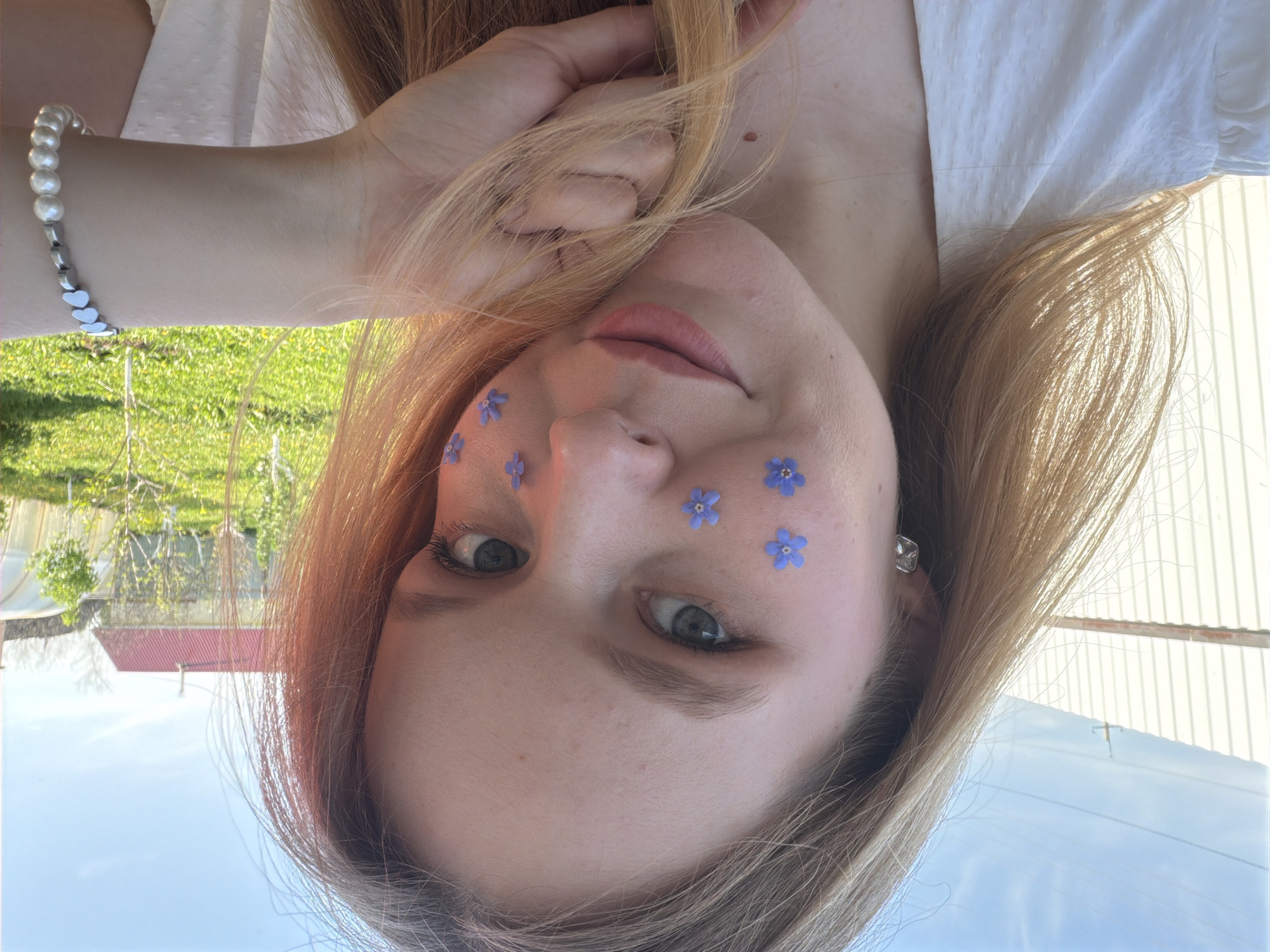 A young woman poses upside down, seemingly outdoors, with several small, light blue flowers lightly adhered to her face around her eyes and nose. She has light skin, light brown or strawberry blond hair, and gray eyes. She is wearing a white top. A bracelet made of pearls and silver-colored heart shapes is visible on her wrist.
In the background, upside down, is greenery suggesting grass, bushes, and trees. A building with a red roof and a white building are visible. Power lines are also visible. The location is likely the city of Кадряково, Russia.