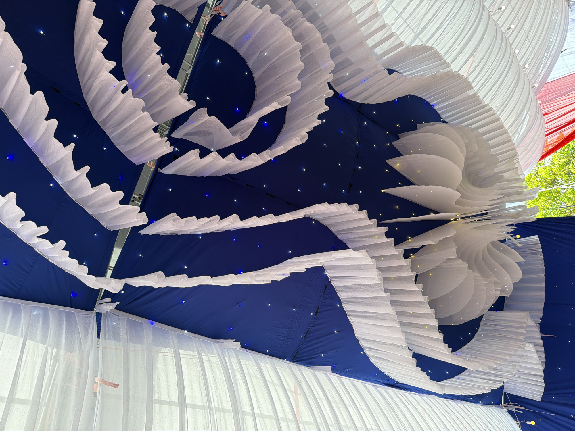The image shows an interior view of what appears to be a decorated tent or canopy, possibly for an event or celebration. The background is a dark blue material dotted with small lights, reminiscent of a night sky. Across this backdrop are draped layers of white fabric, some arranged in wavy, ruffled patterns and others in a more linear, cascading style. The image is taken from a low angle, looking upwards toward the ceiling of the structure. The location is Tay Son Commune, Vietnam.