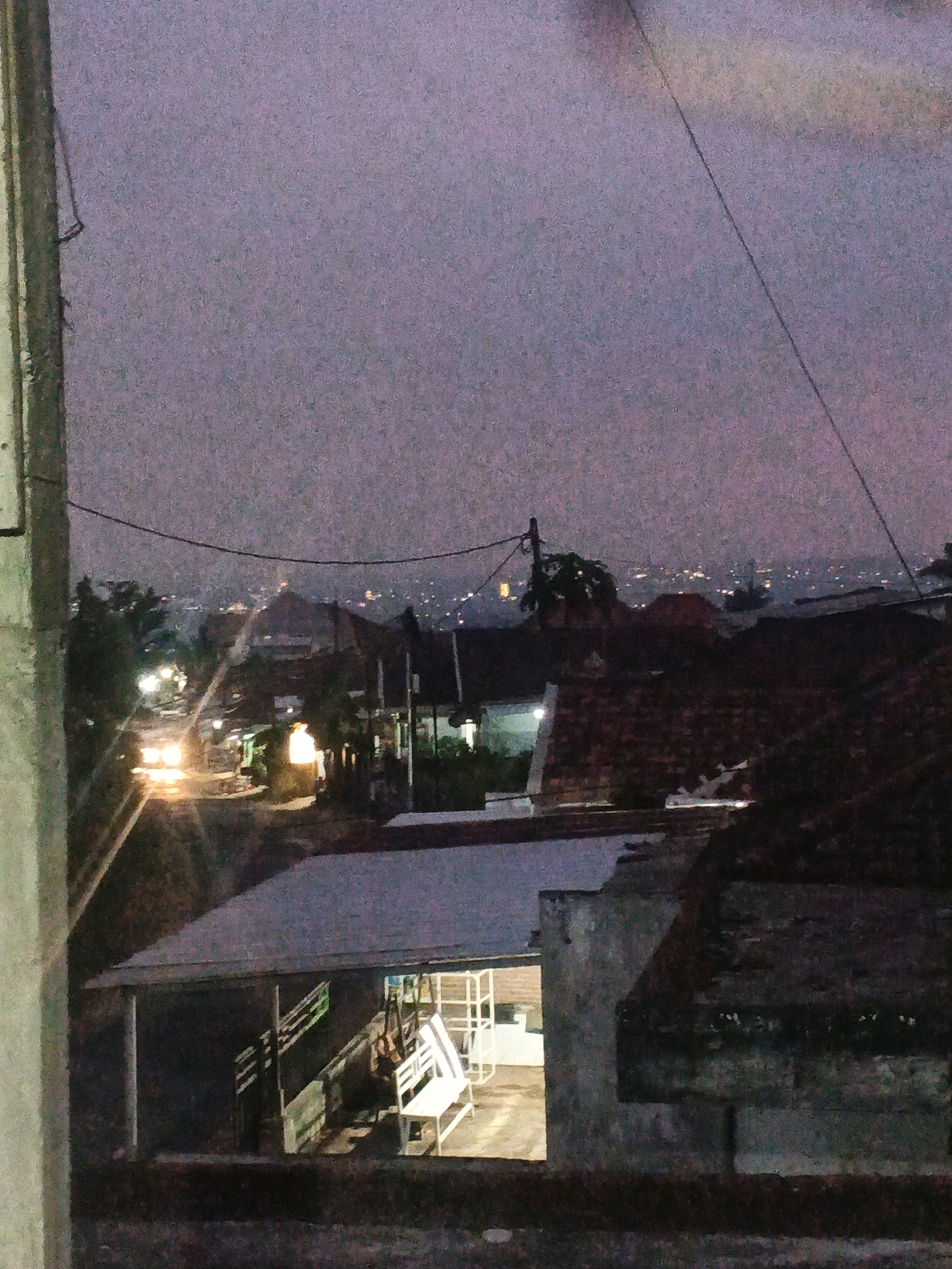 This is a low-light, slightly noisy photograph taken at dusk or dawn, capturing a residential area. In the foreground, a light-colored concrete pillar is visible on the left side. Beyond this, a tiled roof structure is prominent, with a portion of a building featuring a white bench and railing. Further into the mid-ground, several houses with terracotta-colored roofs are clustered together, and a street with illuminated vehicles and streetlights suggests ongoing activity. In the distance, a hazy cityscape with scattered lights is visible under a pale purple sky. The overall impression is of a suburban or semi-urban environment in Bocek, Indonesia, during twilight hours. There are no people clearly visible in the image.