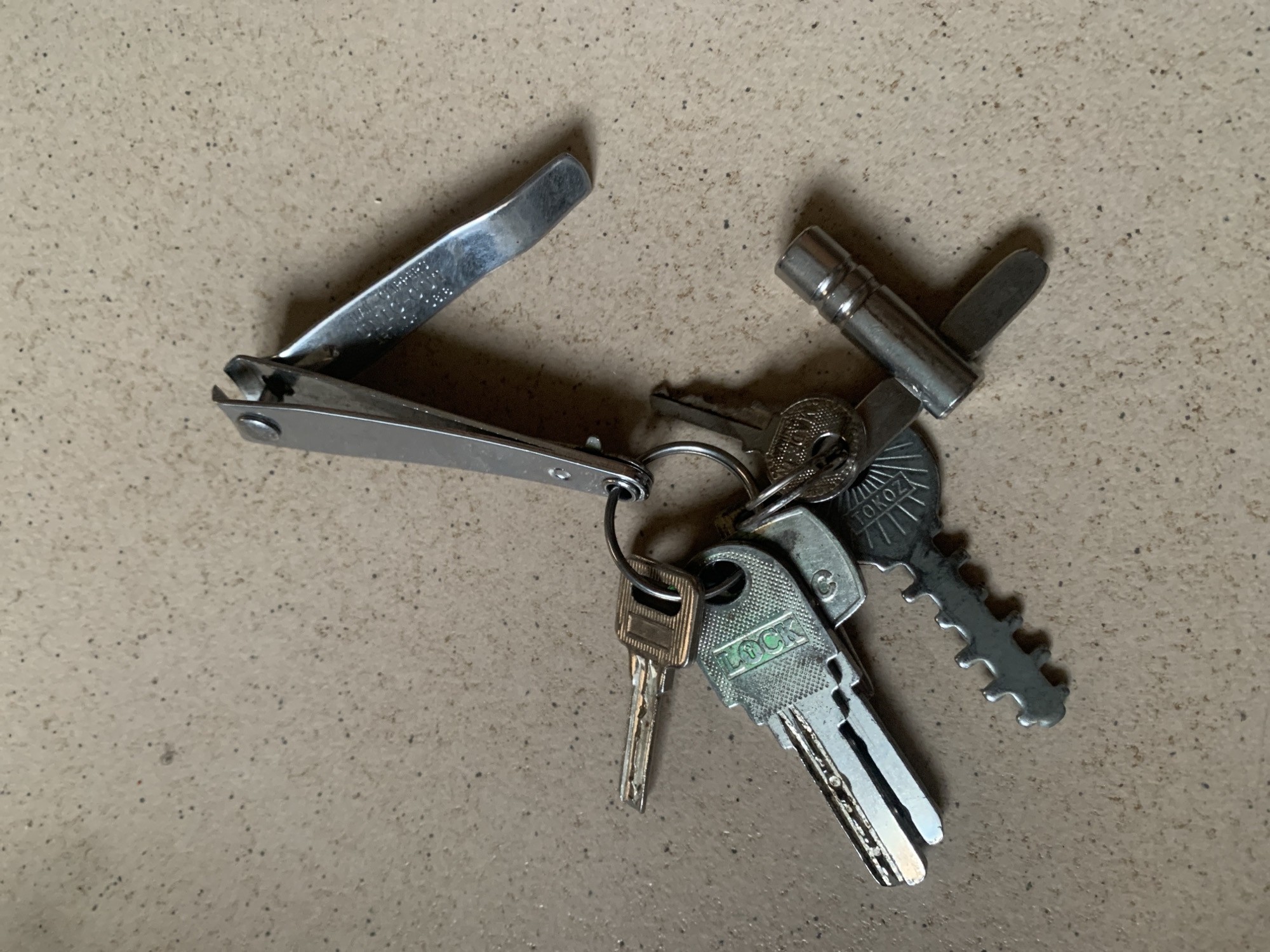 A set of keys and a nail clipper are on a speckled tan surface. The keys are attached to a silver keyring. One key is silver and has a gold stripe. Another key is silver and has "LOCK" printed on it in green. A third key is silver and has "TOKOZ" printed on it. A fourth key is silver and has a ridged edge. A silver nail clipper is partially open and resting on top of the keys.