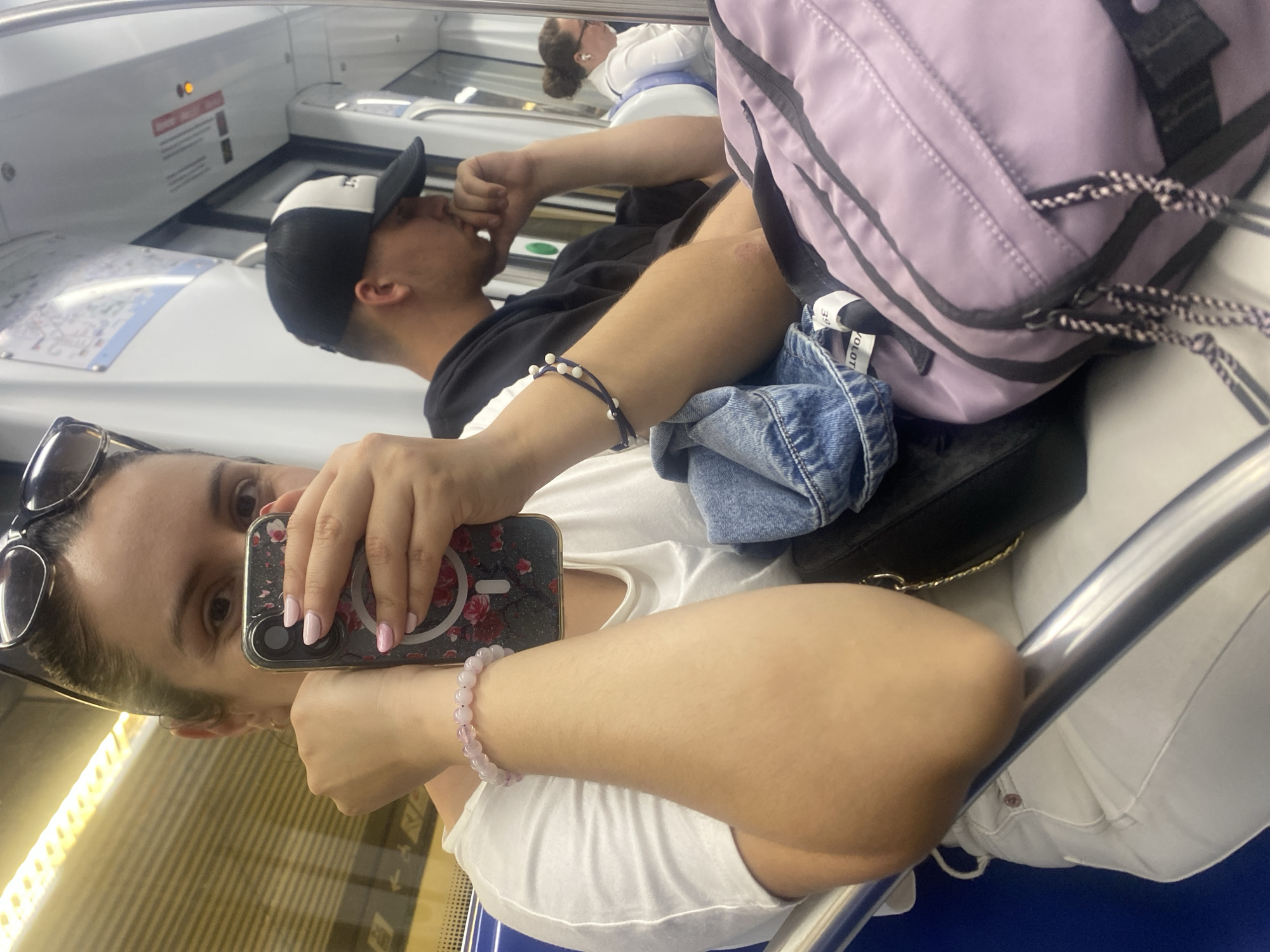 The image depicts the interior of a subway train, likely within the city of l'Hospitalet de Llobregat, Spain, as indicated by the user's request. In the foreground, a woman with sunglasses perched on her head is holding a smartphone, its floral-patterned case facing the camera. She wears a white t-shirt and a light-colored bracelet on her left wrist. Her right arm is bent, with a dark purse and a pair of denim jeans visible near her. Beside her, a man wearing a black and white baseball cap is seated, looking downwards with his hand near his chin. He is wearing a dark hoodie. A light purple backpack with black trim and decorative pull cords is placed on the seat in the foreground. In the background, another woman with her hair in a bun is visible, wearing a white shirt and earbuds. The train's interior features metallic poles and seating. The lighting suggests an interior environment, possibly during daylight hours.