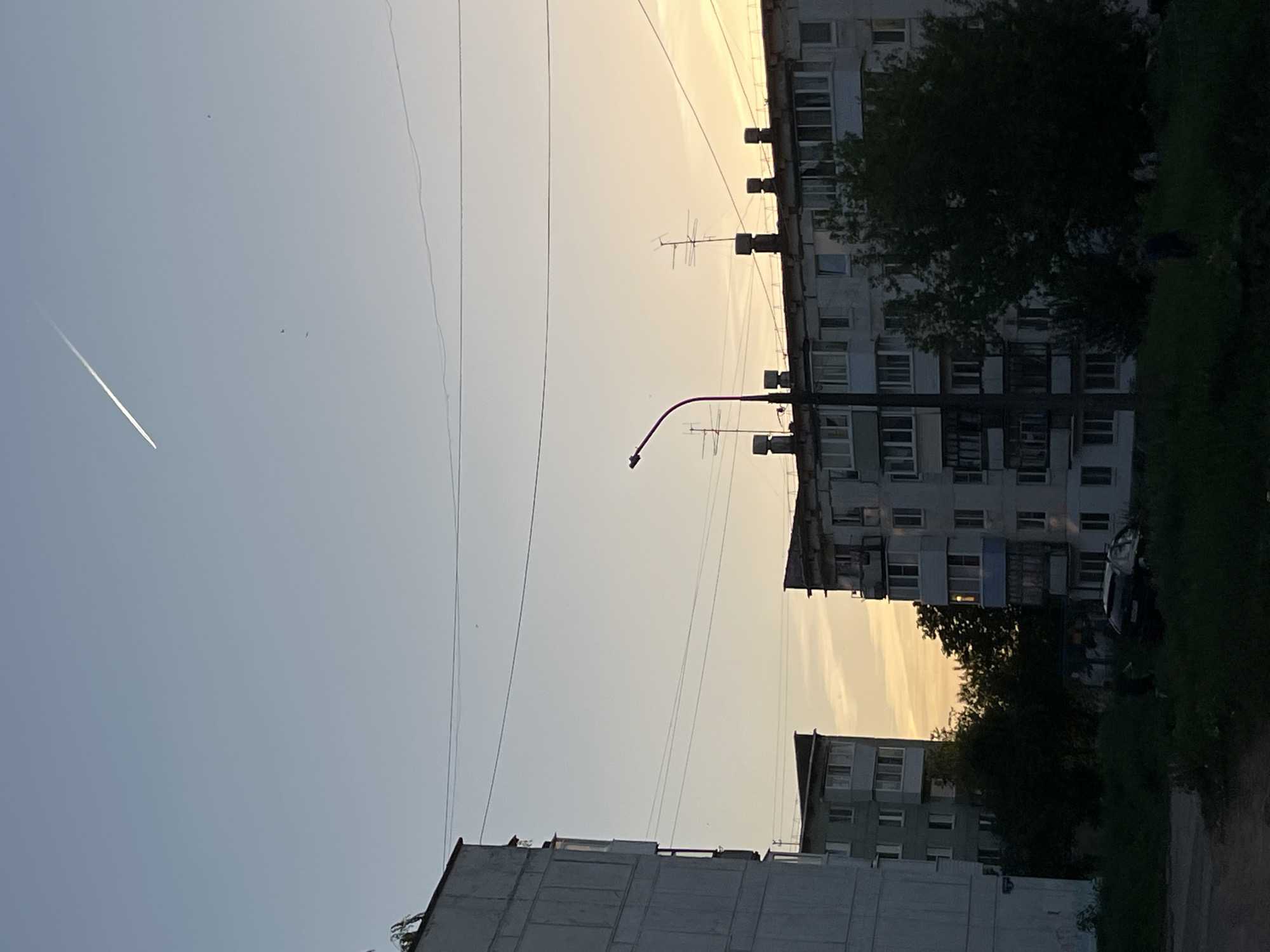 The image shows a view of a typical residential area in the city of Chaikovsky, Russia. In the foreground and slightly to the left, there is a concrete building with a flat roof. Overhead, there are several power lines stretching across the sky. To the right, a taller apartment building is visible, with numerous windows and balconies. A tall lamppost stands in front of the apartment building, with a curved arm extending outwards. Trees and grass grow along the right edge of the image, suggesting a green space or park area. In the upper-left corner of the image, there is a faint contrail from an airplane in the sky, indicating recent air traffic. The sky has a slightly yellowish hue, suggesting it might be either early morning or late evening. The overall scene captures a mundane, everyday view of urban life in Russia.