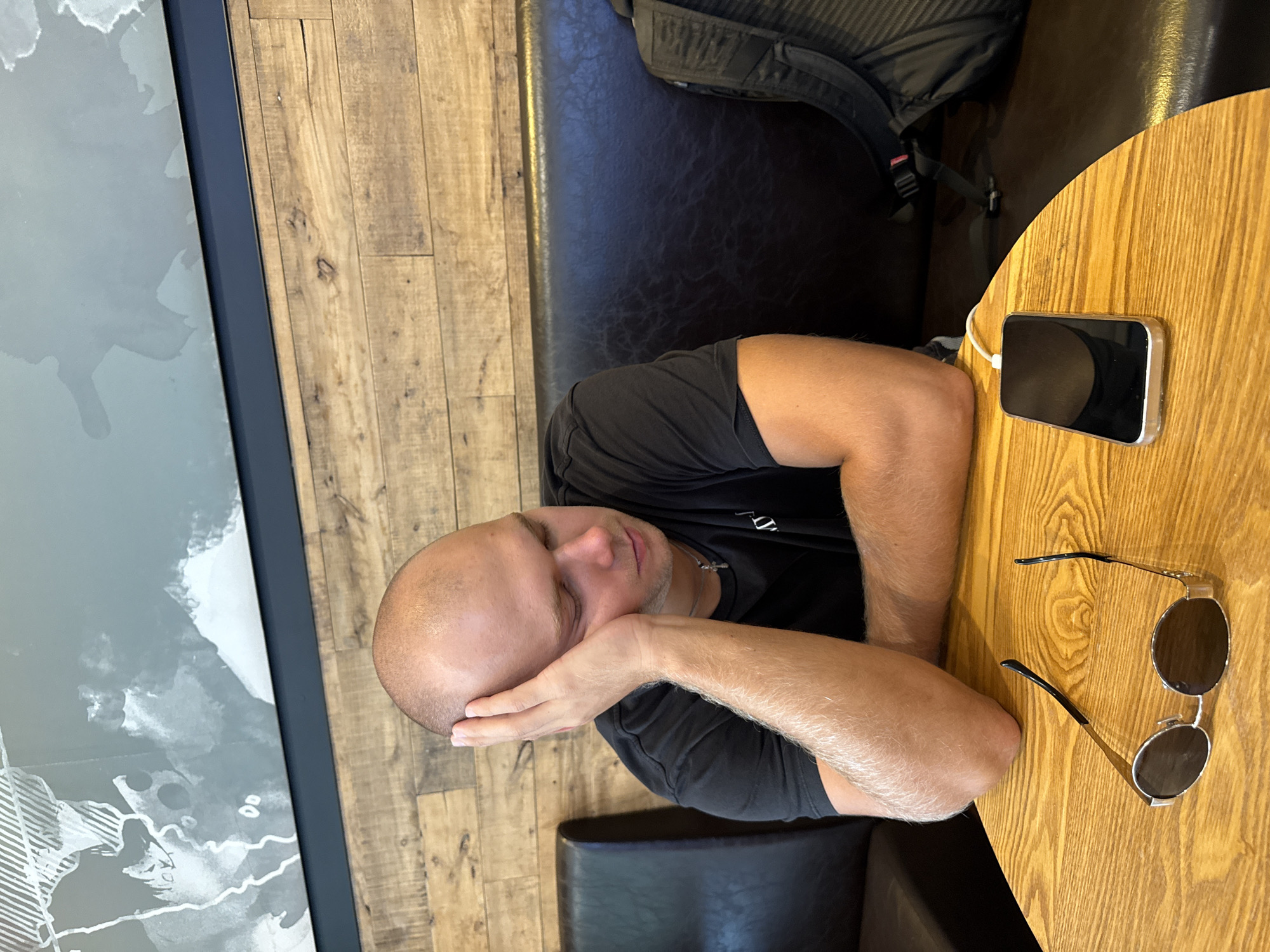 A bald man in a black t-shirt rests his head on his hand, eyes closed, appearing to be asleep or resting. He is sitting at a round wooden table in what appears to be a cafe. On the table in front of him, there are a pair of sunglasses and a smartphone connected to a charger. Behind him, a dark brown leather booth seat is visible, with a black backpack placed on it. The wall behind the booth is paneled with light brown wood, and a large abstract mural with grey and white tones is mounted above it. This scene is consistent with a cafe located in Tianhe District, China, given the typical cafe decor and urban setting.
