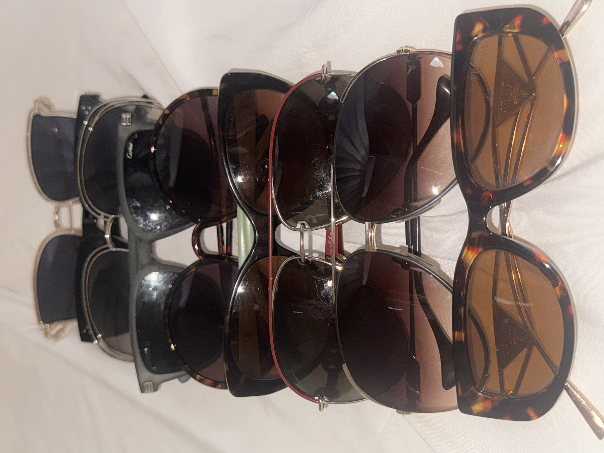The image shows a row of sunglasses displayed on a rack, likely for sale. There are at least six pairs of sunglasses. The brands visible include Cartier, Tiffany & Co., and Prada. The backgrounds of the glasses include various colors such as white, black, and brown. The location is unknown, and it is impossible to tell it is Davao City, Philippines, from the image.