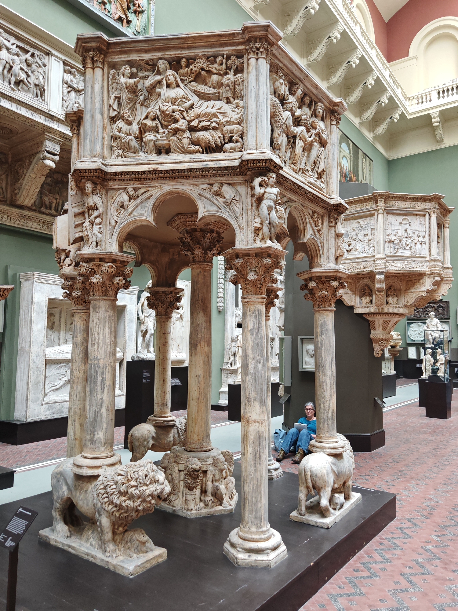 The image shows a detailed, intricately carved marble pulpit within a museum setting, likely the Victoria and Albert Museum in London, given the collection and architectural style. The pulpit rests on several pillars, each supported by sculpted lion figures at the base. The top of the pulpit is adorned with relief carvings depicting biblical scenes. A woman sits on the floor behind one of the pulpit's pillars, reading from a tablet. The museum environment includes other sculptures and artwork in the background, with a patterned brick floor and high ceilings.